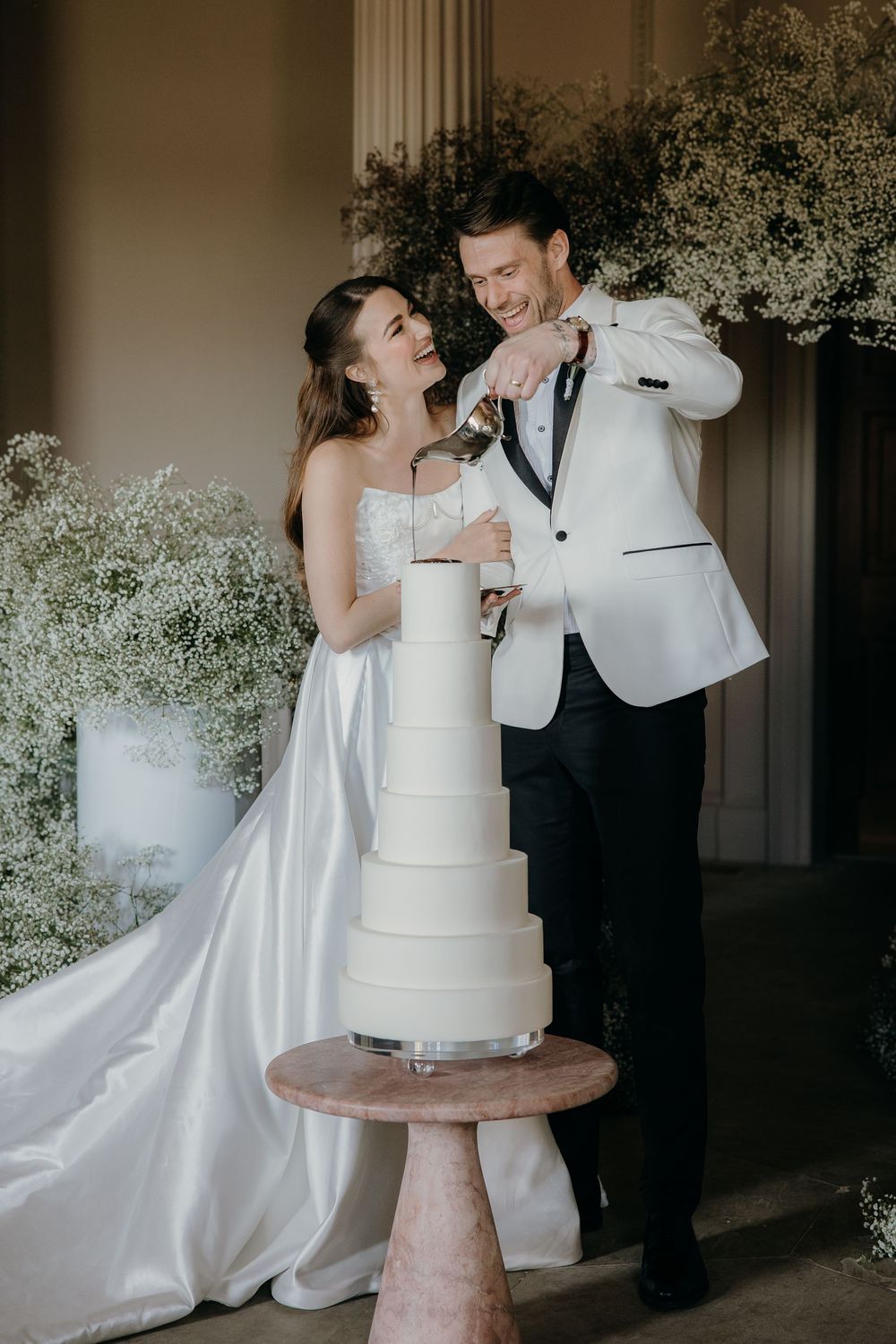Bride and groom pouring melted chocolate onto a white tiered wedding cake, surrounded by baby's breath floral arrangements at Denton Hall.