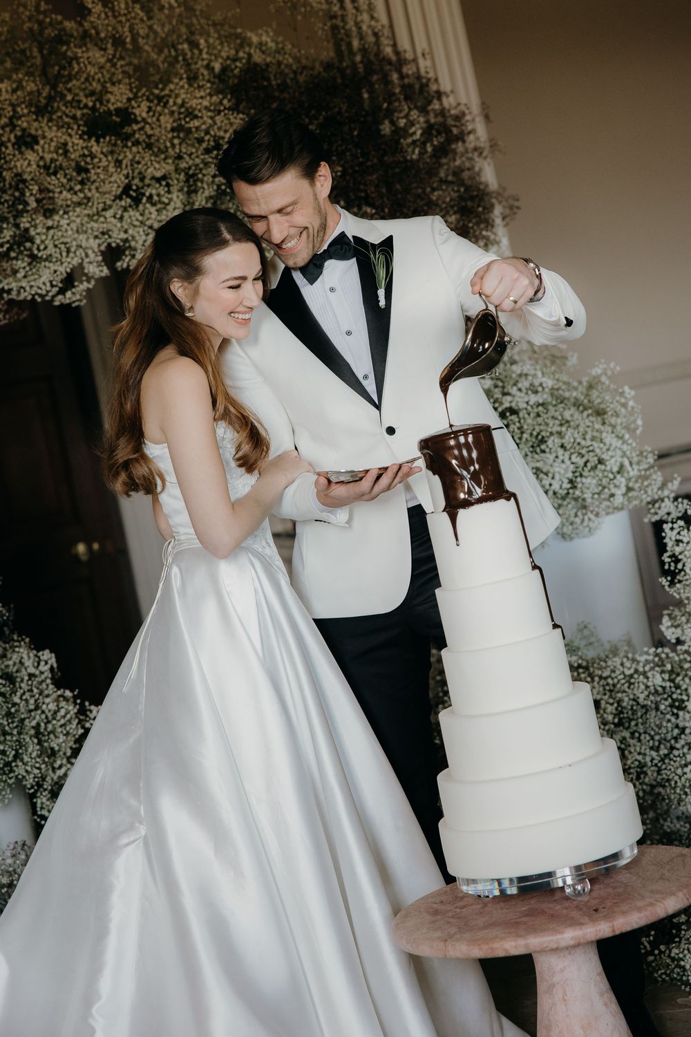 Bride and groom pouring melted chocolate onto a white tiered wedding cake, surrounded by baby's breath floral arrangements at Denton Hall.