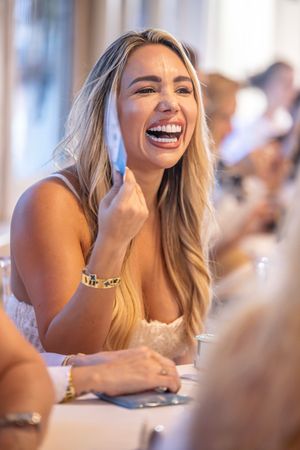 A laughing blonde woman in a white dress enjoys a lively conversation at a restaurant table, wearing gold jewelry.