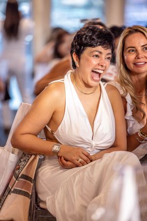 Two women in white dresses laughing joyfully at an indoor event, one with short dark hair wearing a gold necklace.