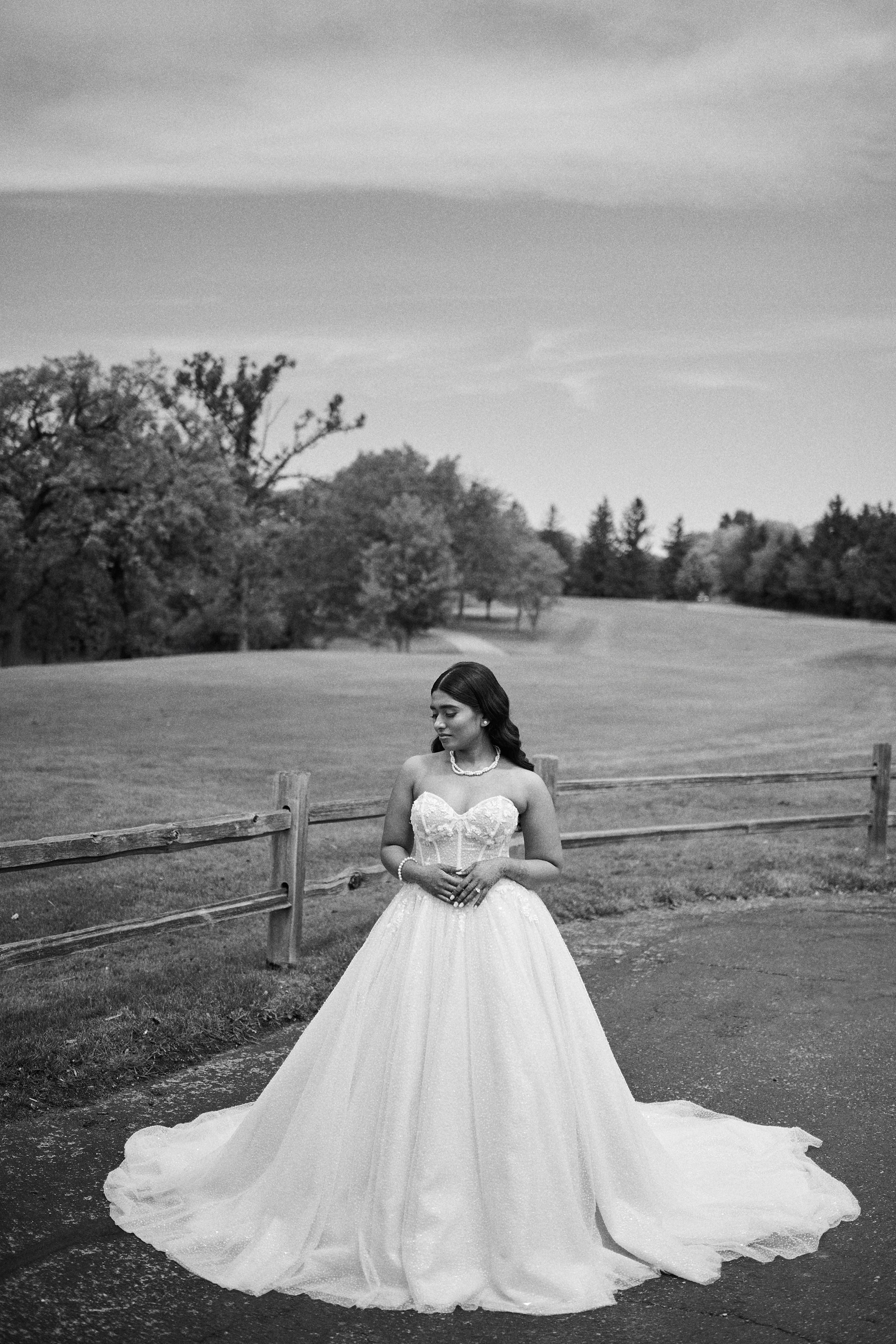 Bride in strapless ball gown wedding dress poses outdoors near rustic fence with rolling green hills in black and white photo.