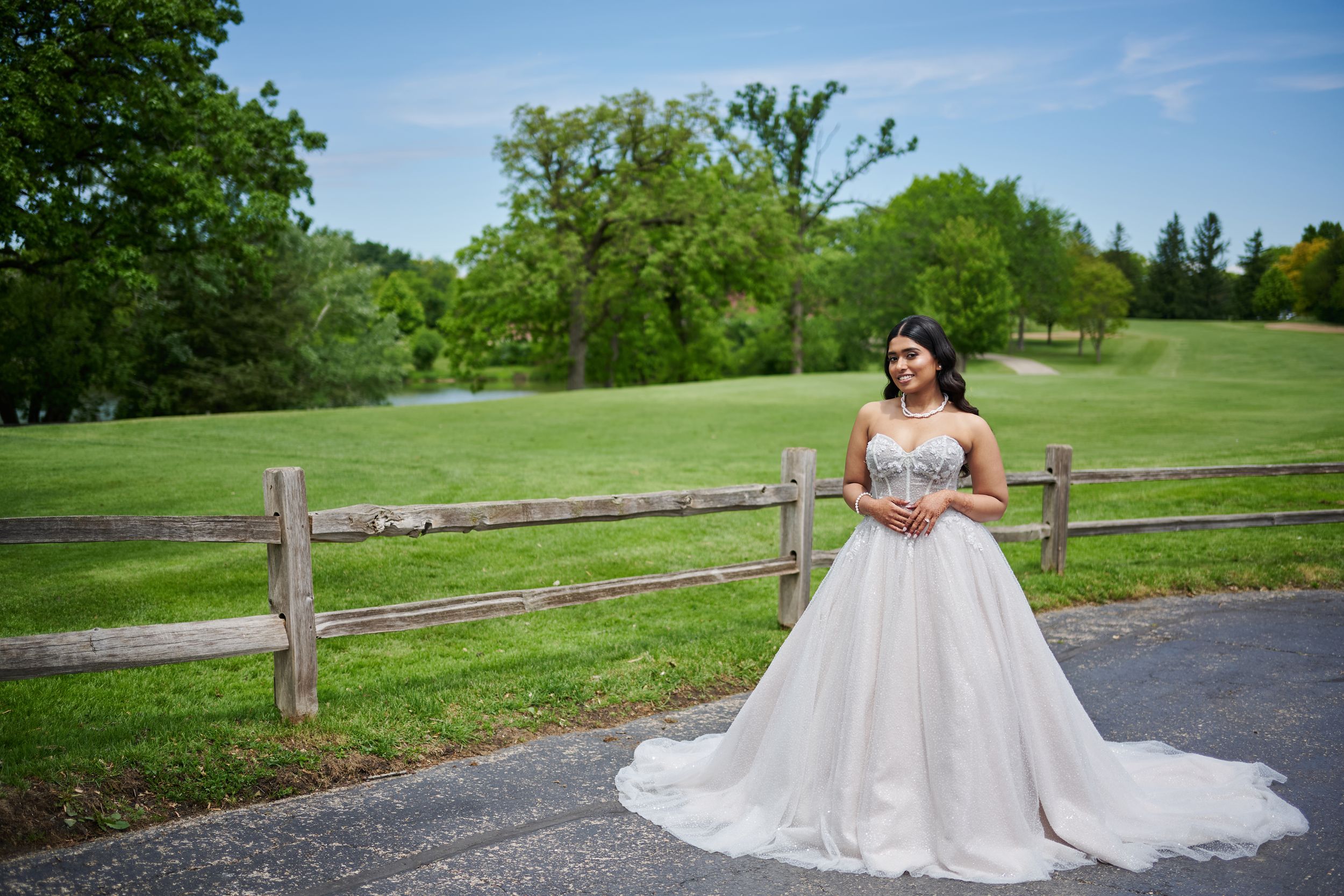Bride in elegant ballgown with beaded bodice poses by rustic fence on lush green golf course under blue sky.