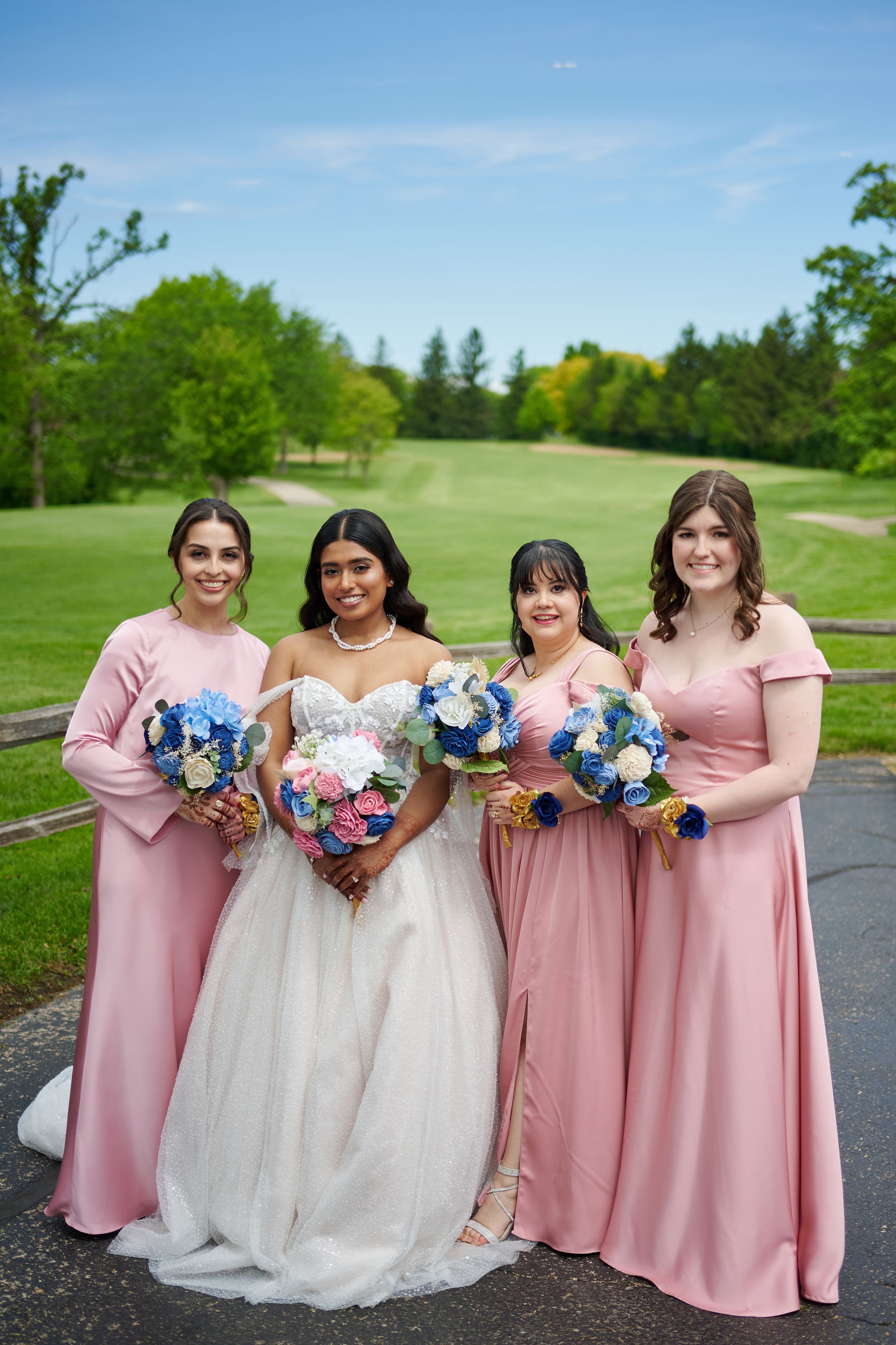 Bride in white gown poses with three bridesmaids in pink dresses holding floral bouquets on a green golf course.