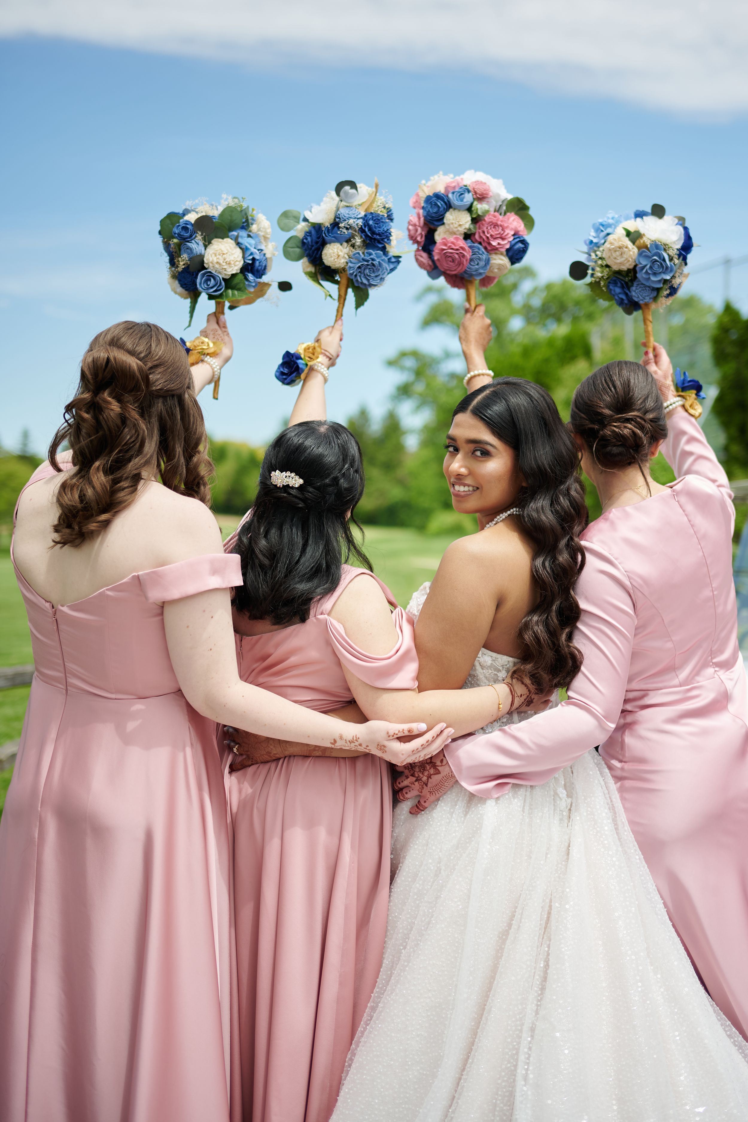 Bride in white gown with bridesmaids in pink dresses raising floral bouquets outdoors on a sunny day.