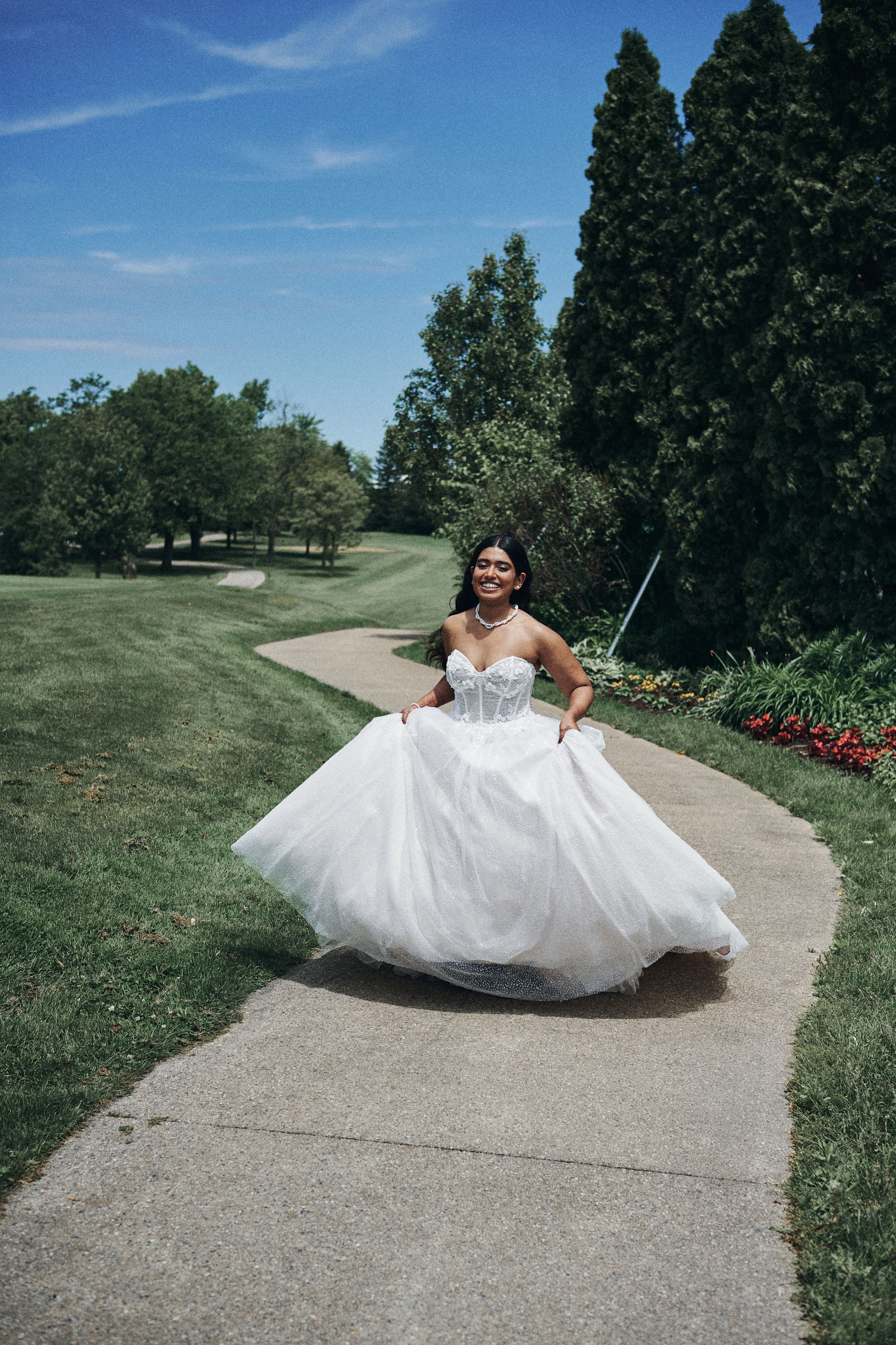 Bride in a flowing white ballgown twirling on a garden path, smiling joyfully on a sunny day.