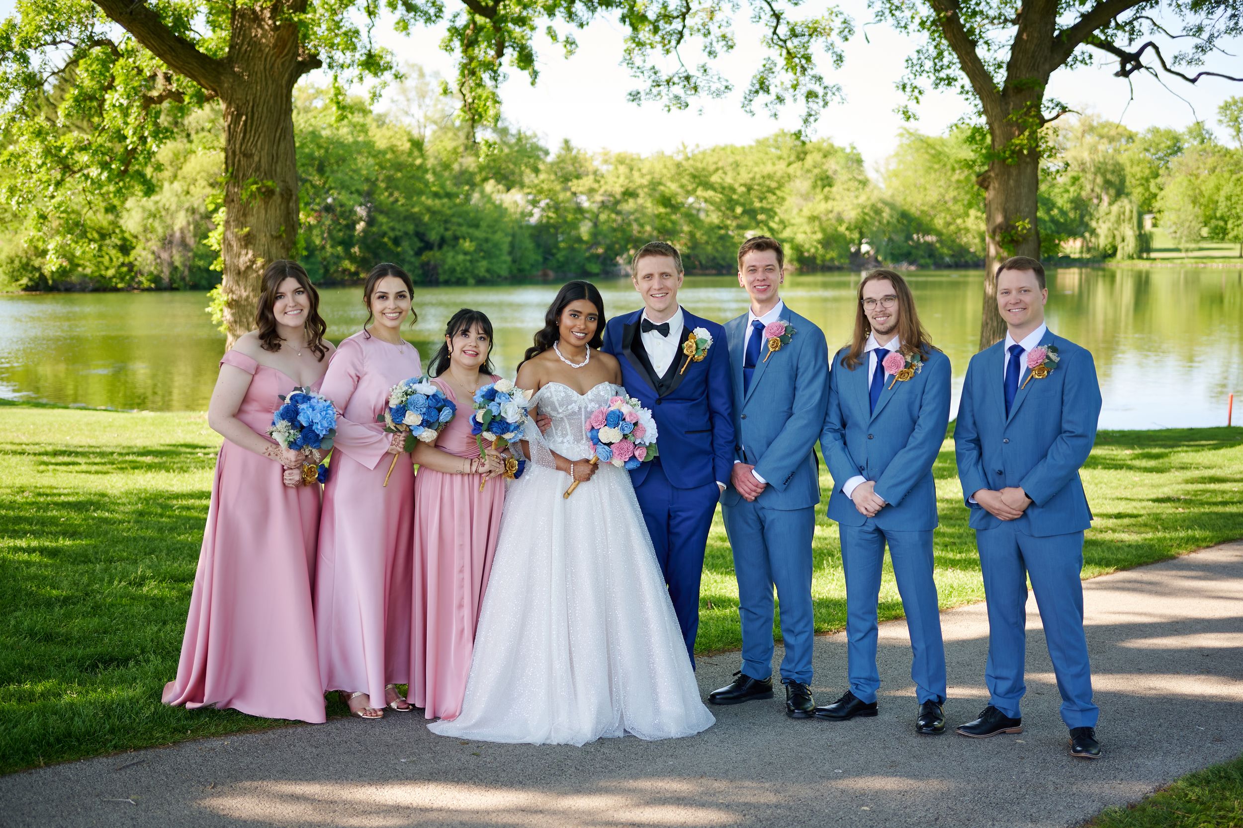 Wedding party poses outdoors near a pond, bride in white gown with pink and blue bouquet, bridesmaids in pink, groomsmen in blue.