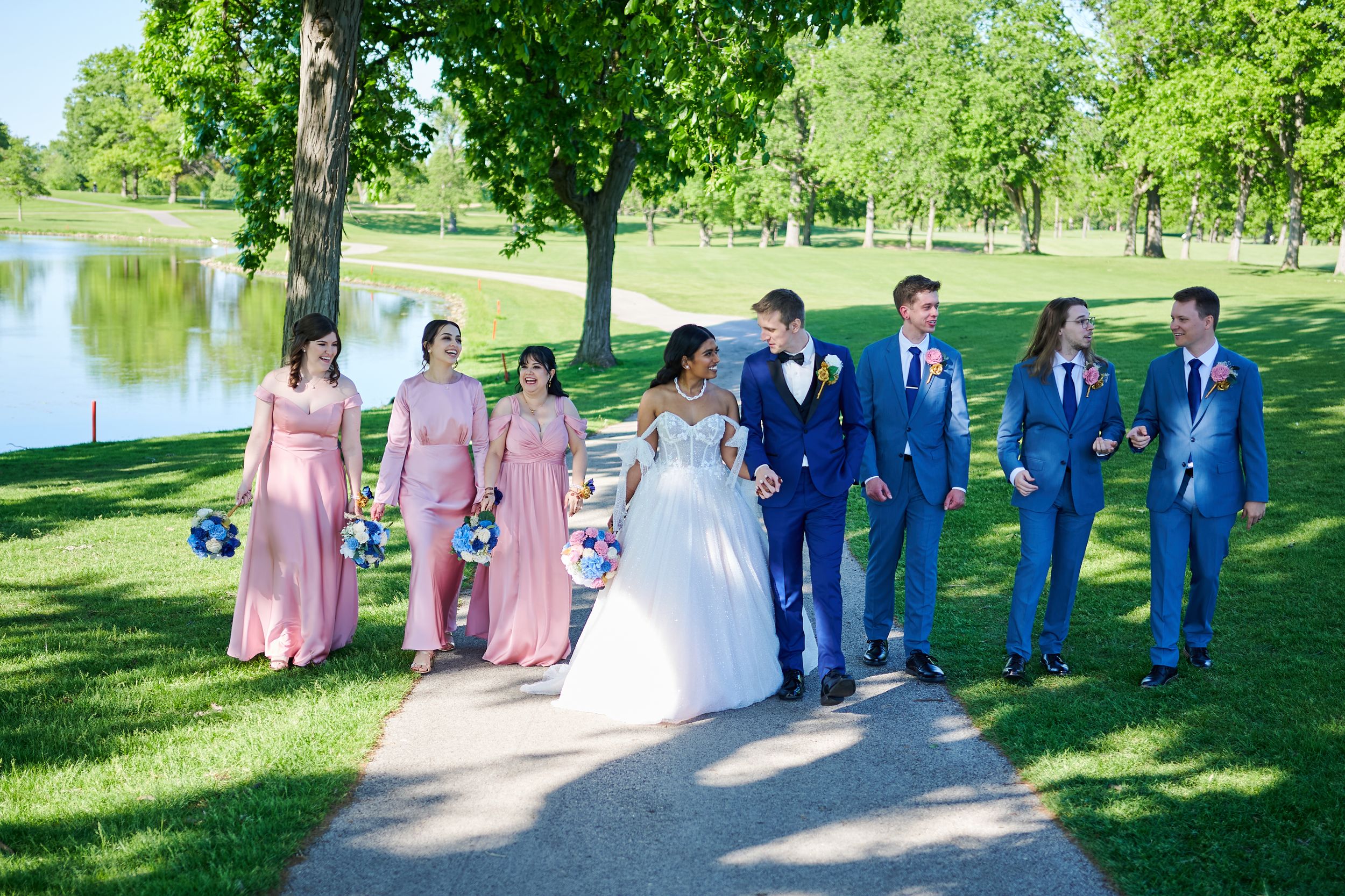 Wedding party walks outdoors near a pond, bridesmaids in pink gowns and groomsmen in blue suits with bride and groom.