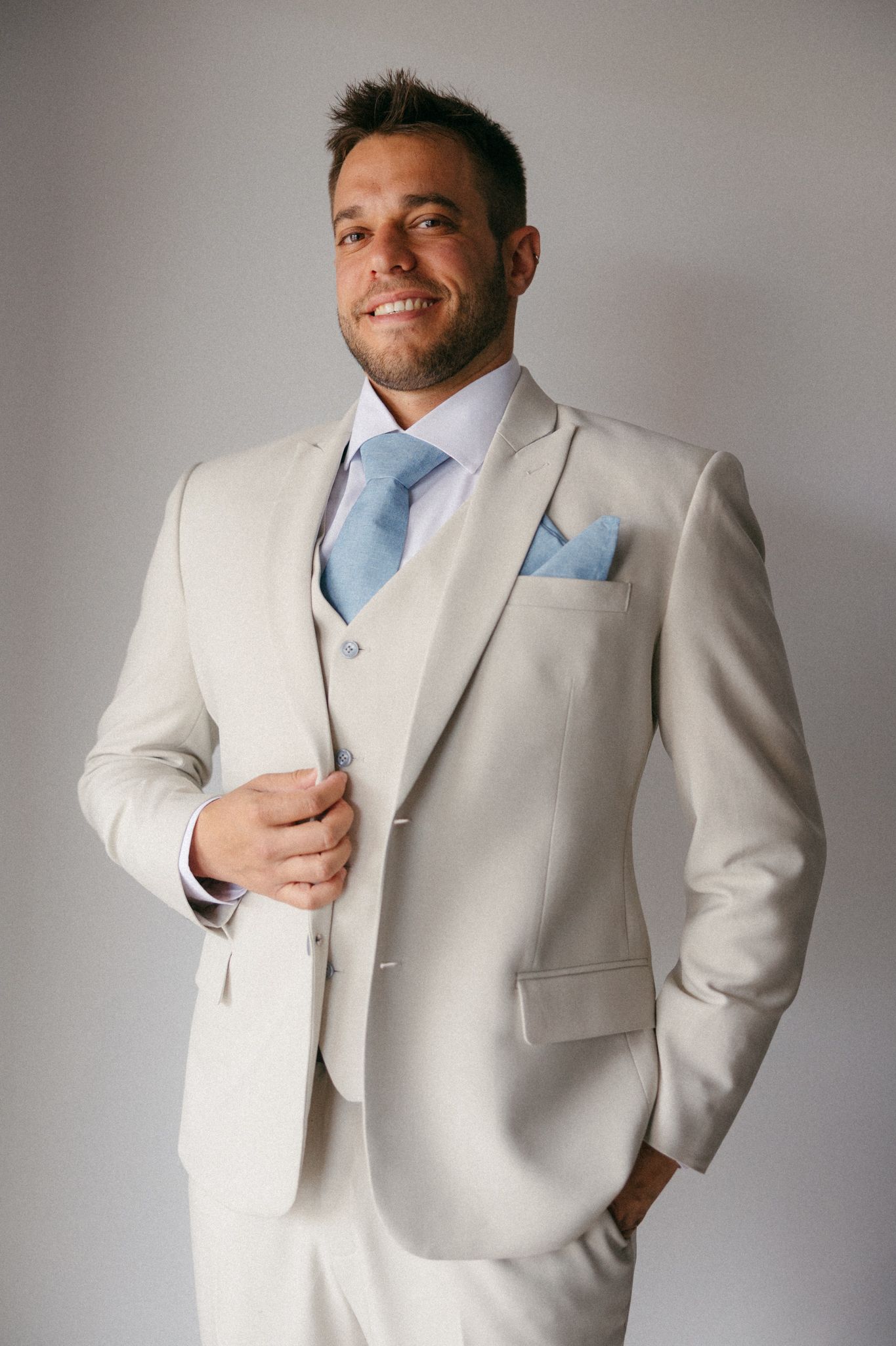 Smiling man in cream three-piece suit with light blue tie and pocket square poses confidently against gray background.