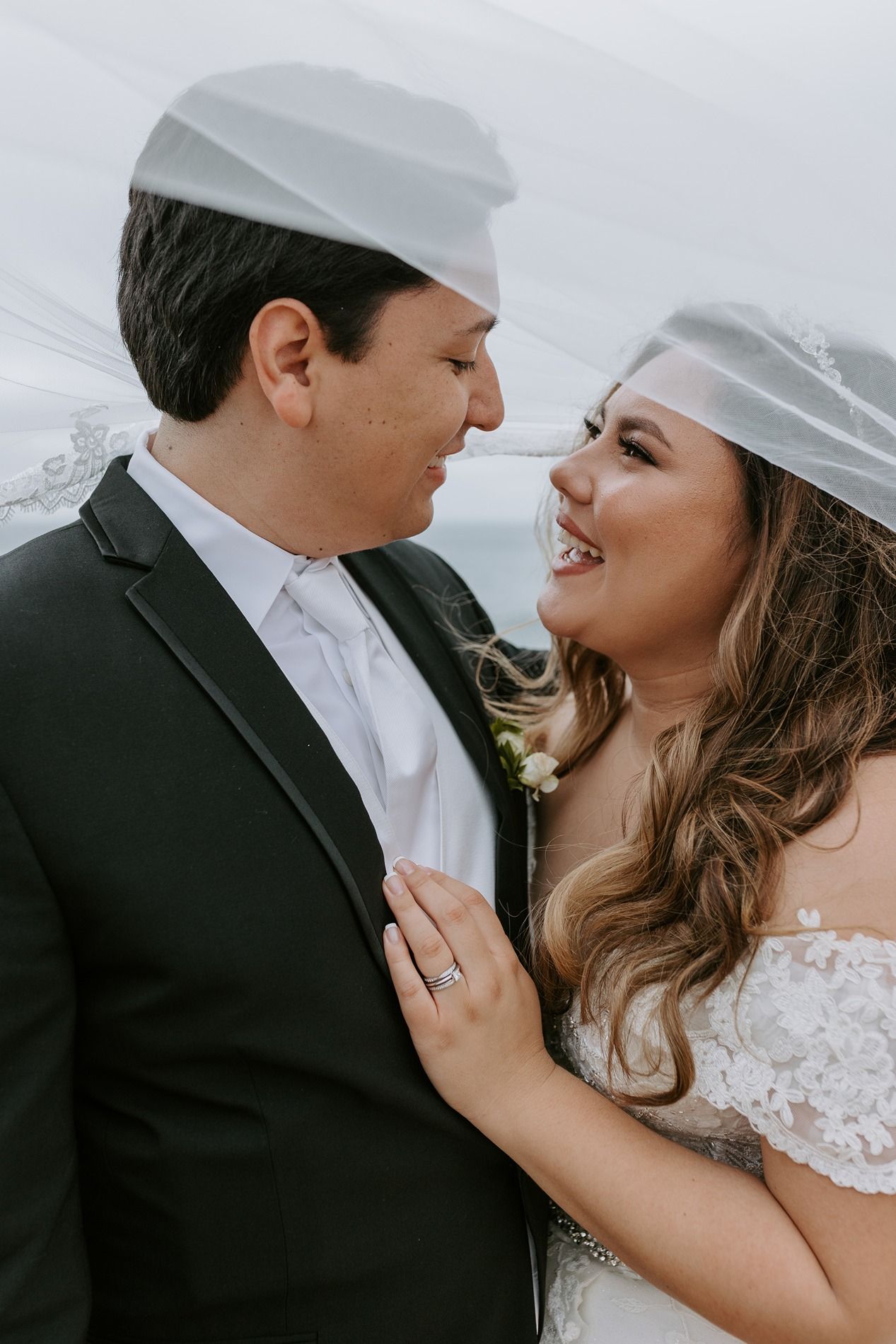 Bride and groom share a romantic moment under a veil, smiling at each other in wedding attire.