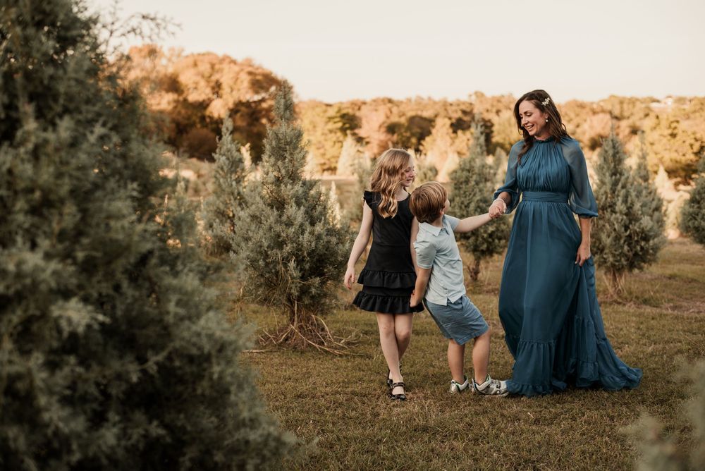 A pregnant woman in a teal gown holds hands with two children in a Christmas tree farm at golden hour.