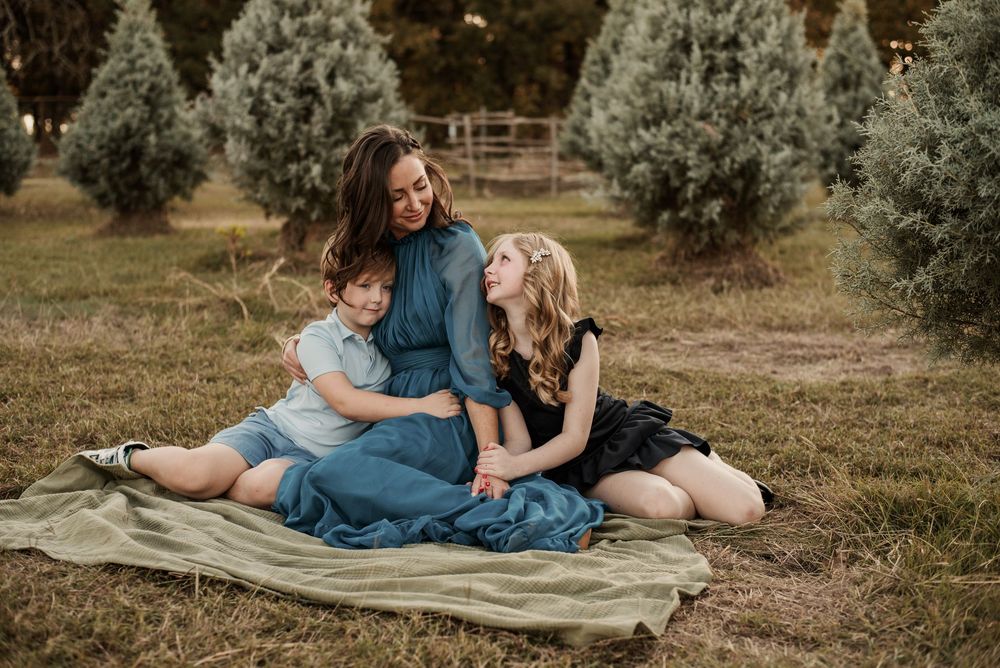 A mother and two daughters sit together on a blanket at a Christmas tree farm, smiling warmly in coordinated outfits.