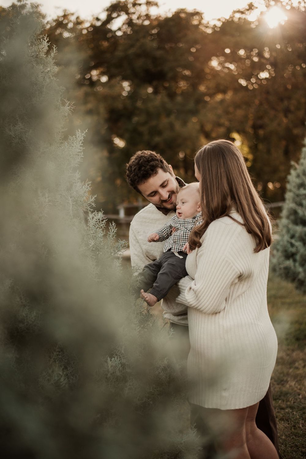 A young family with a baby poses among lush green trees during a warm golden hour outdoor photo session.