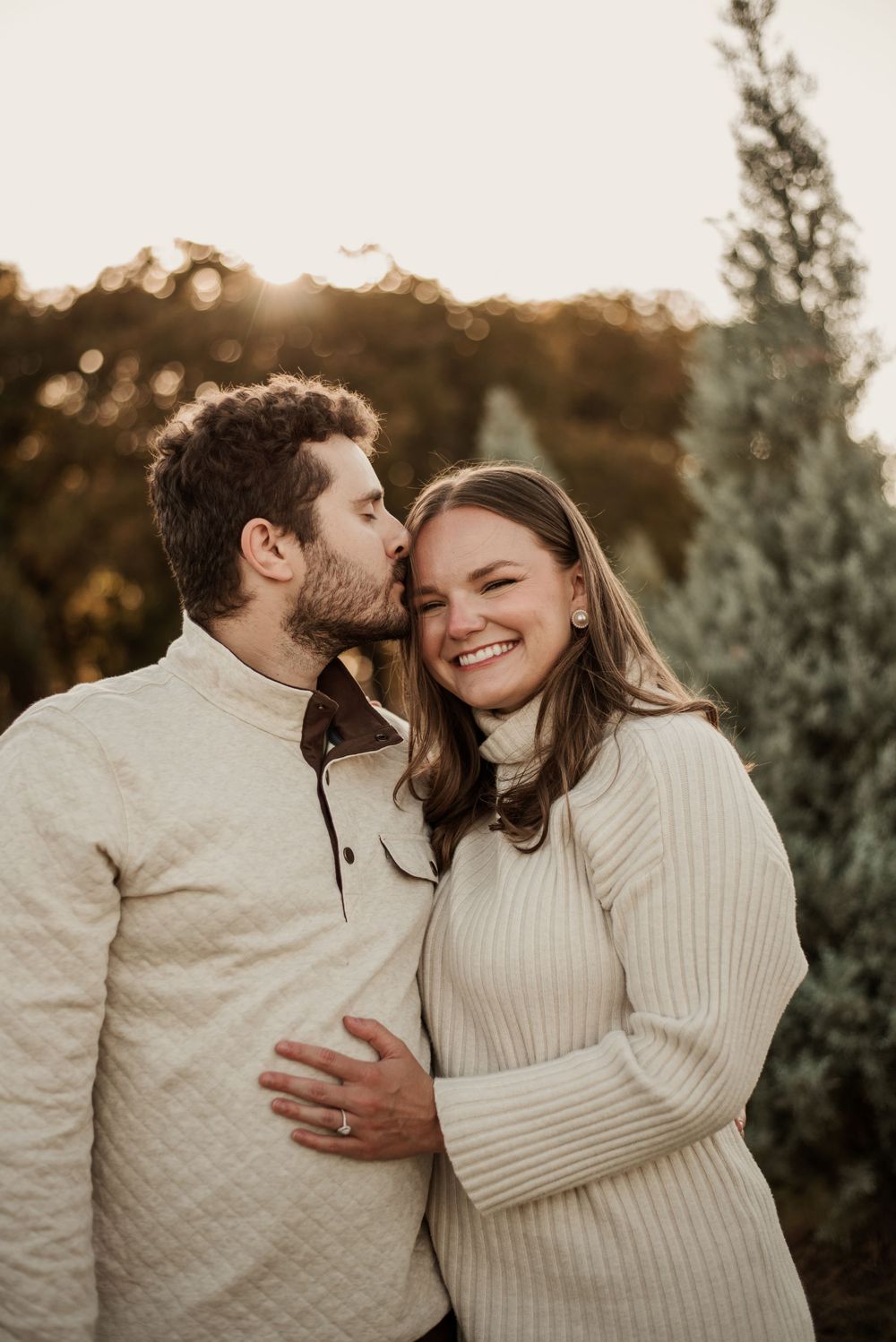 A couple in matching cream sweaters shares a tender kiss at golden hour near evergreen trees.