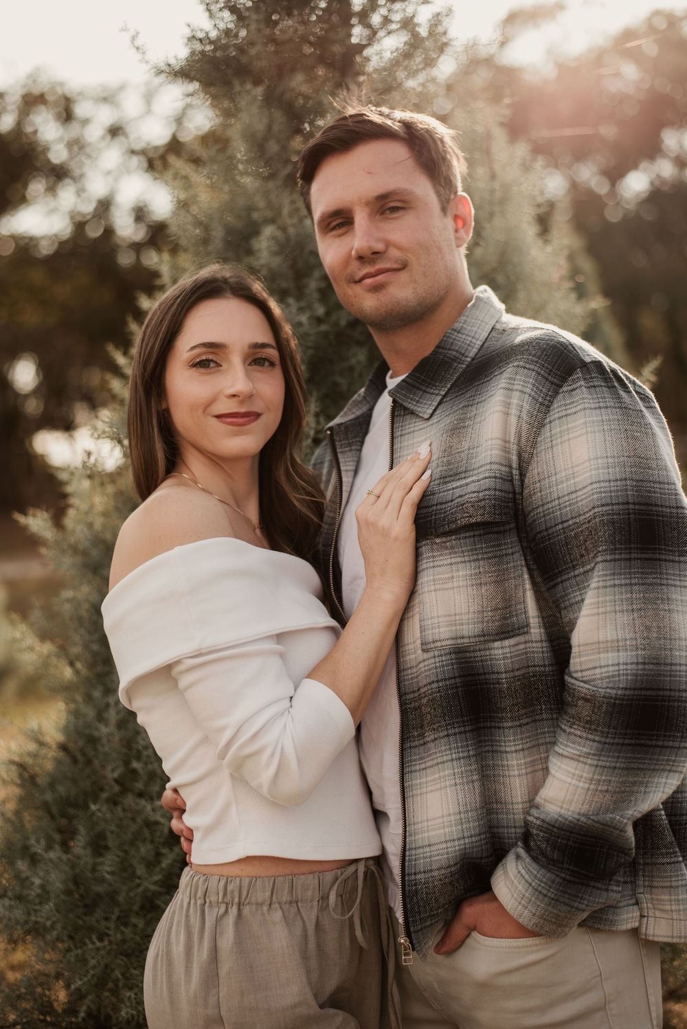 A couple poses outdoors at golden hour, woman in white off-shoulder top and man in plaid shirt, surrounded by trees.
