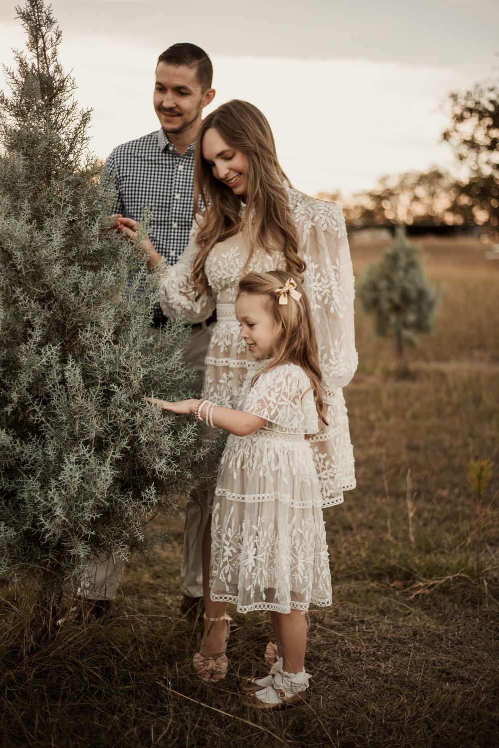 Family of three posing near a pine tree at sunset, mother and daughter in matching lace dresses, father in plaid shirt.
