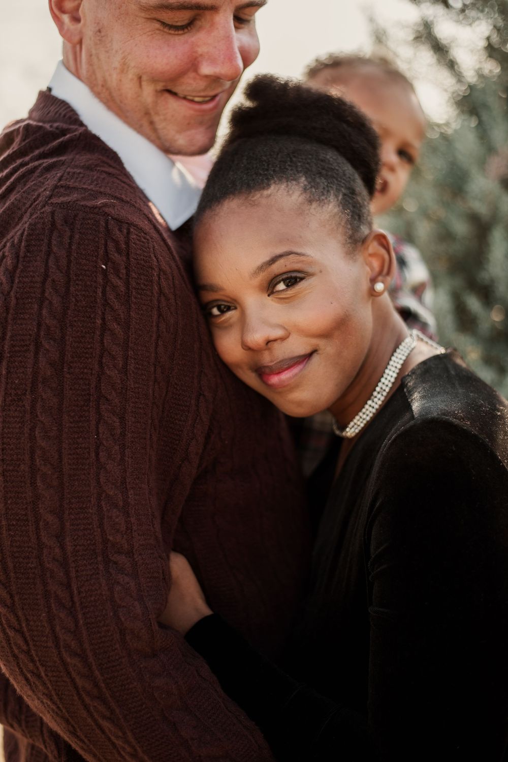 A woman in a pearl necklace smiles while embracing a man in a brown cable-knit sweater outdoors.