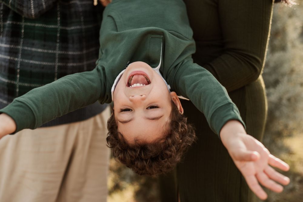 A laughing child hangs upside down with arms spread wide, held by adults in a joyful outdoor family moment.