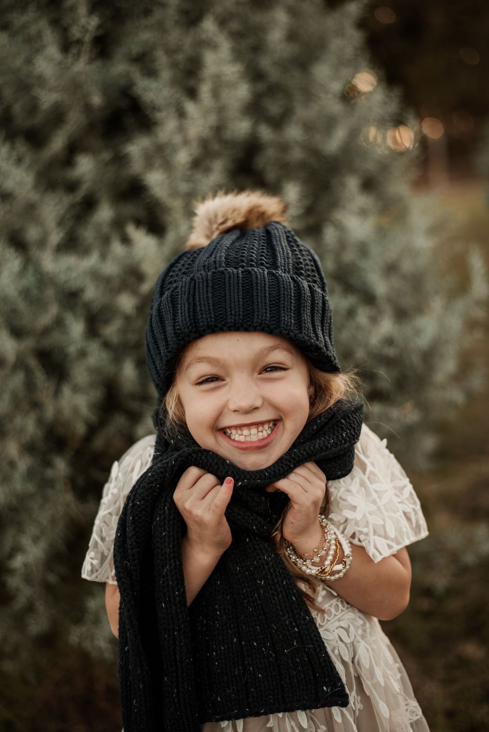 Smiling young girl wearing a black knit beanie with pom-pom and matching scarf, standing near evergreen trees.
