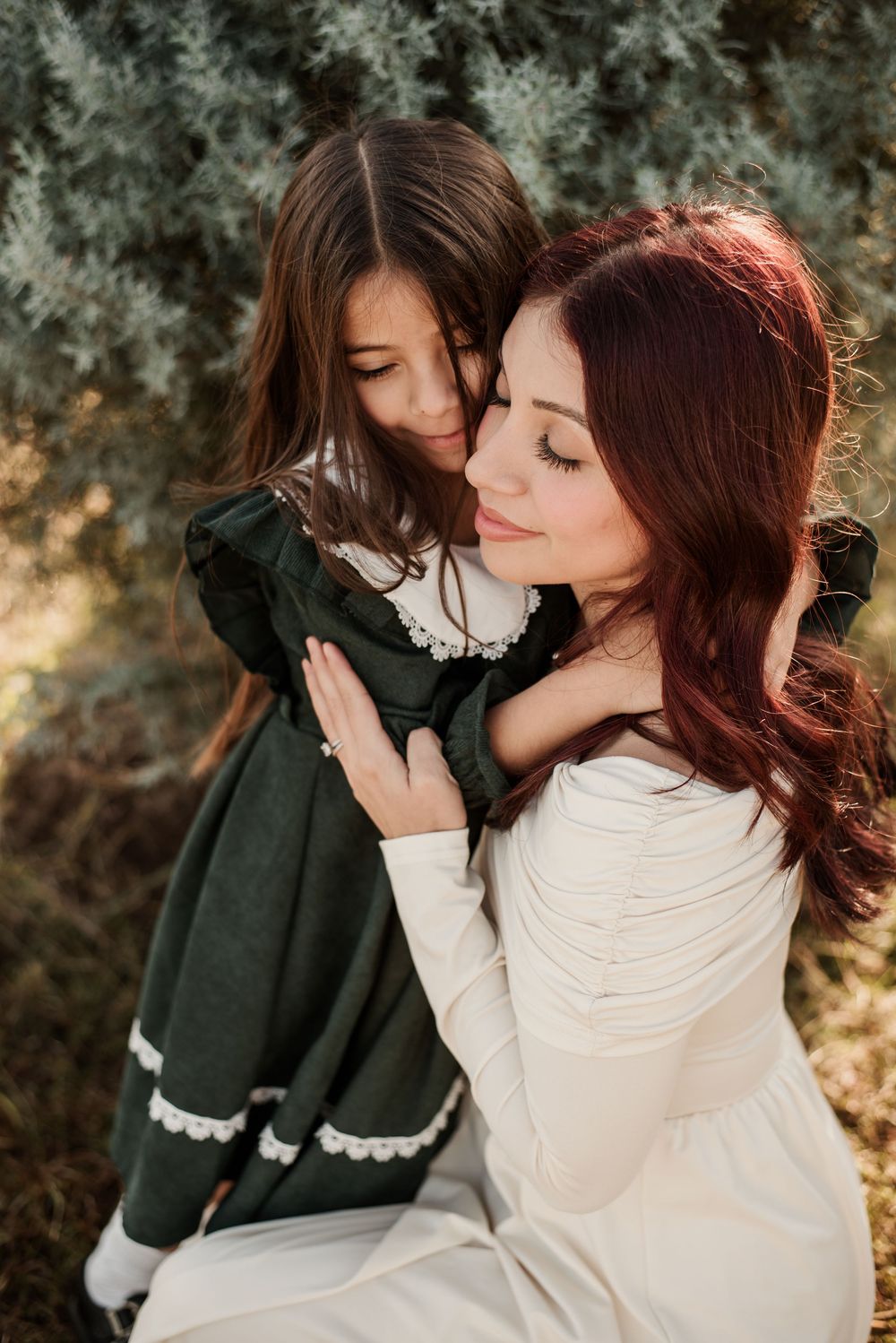 A mother with red hair in a white dress tenderly embraces her young daughter in a dark green dress outdoors.