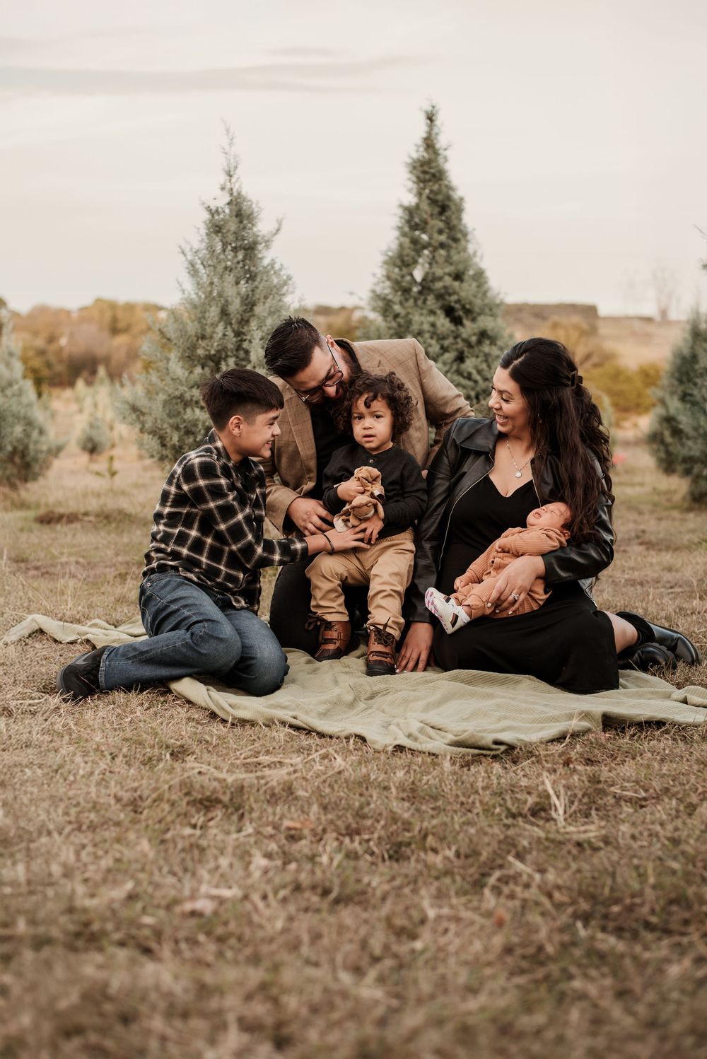 Family of five sits on a blanket at a Christmas tree farm, parents smiling at toddler while holding newborn baby.