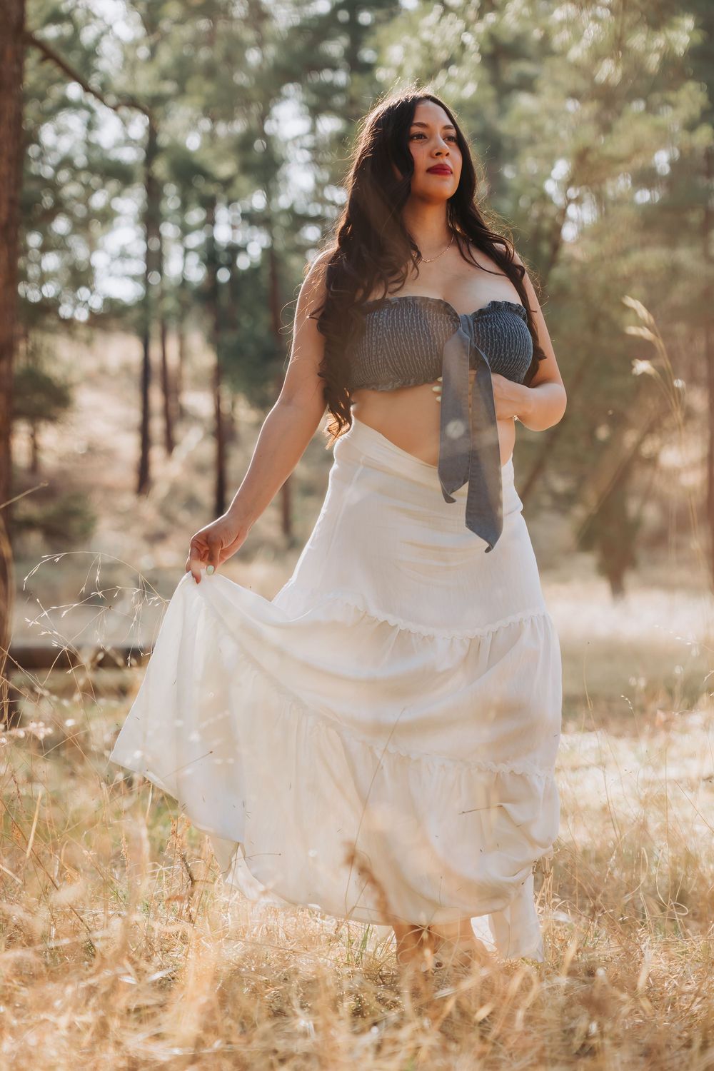 Woman in crop top and flowing white skirt posing in sunlit forest.