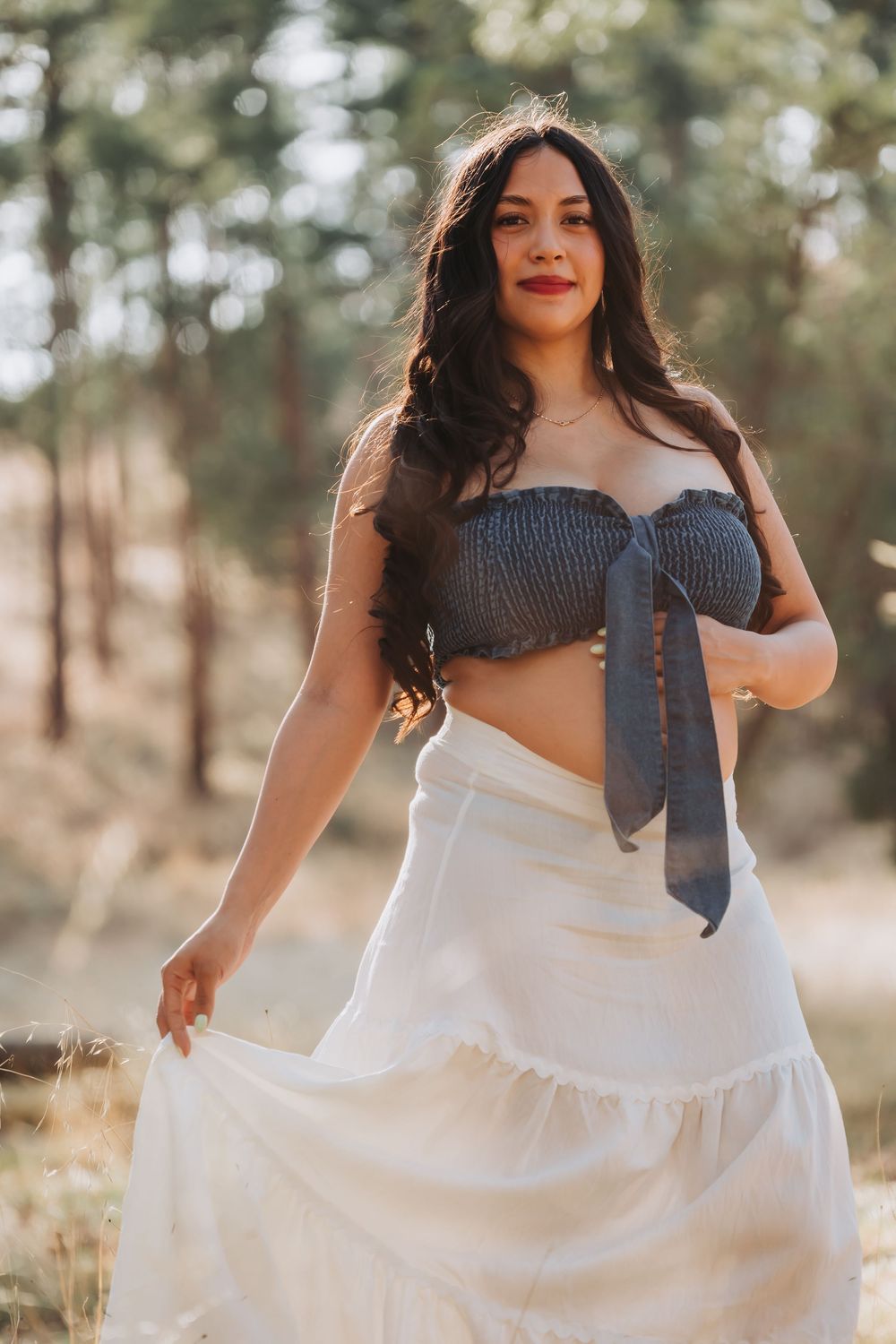 Woman in gray smocked crop top and white maxi skirt posing outdoors in a sunlit forest setting.
