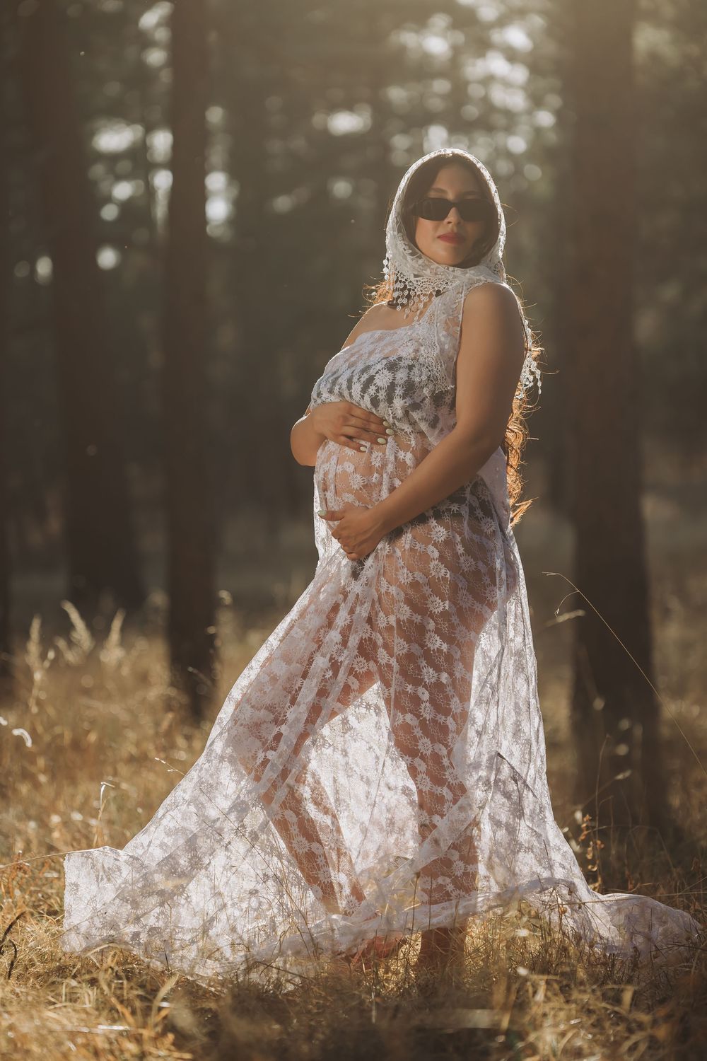 Pregnant woman in white lace dress holding belly in sunlit forest, wearing sunglasses and head scarf.