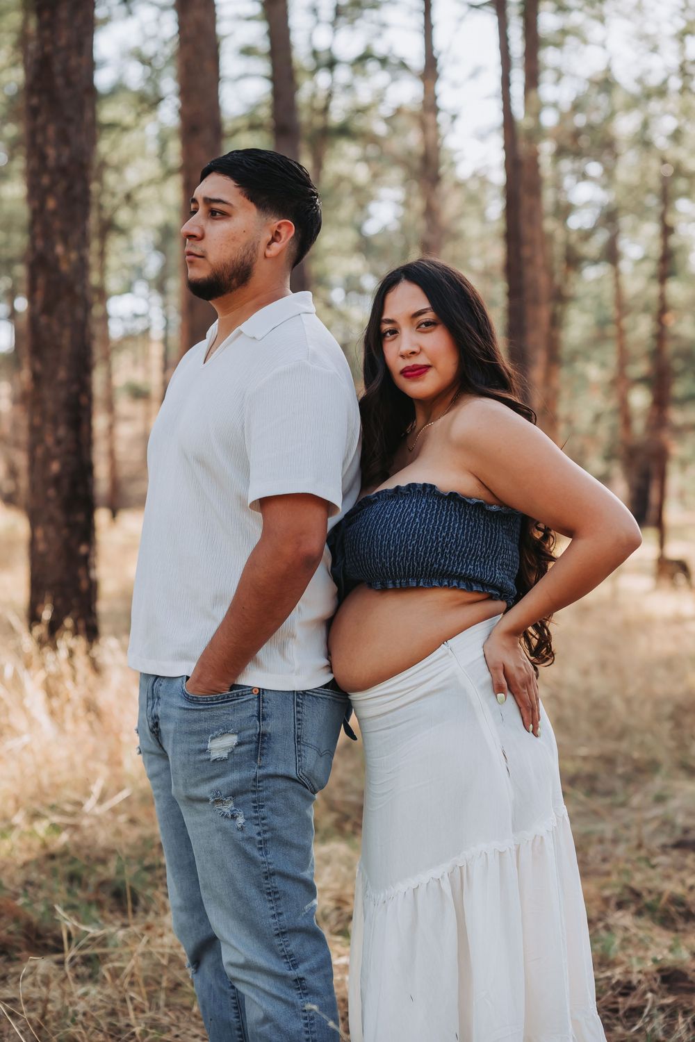 Couple posing in pine forest, man in white shirt and jeans, pregnant woman in navy crop top and white skirt.