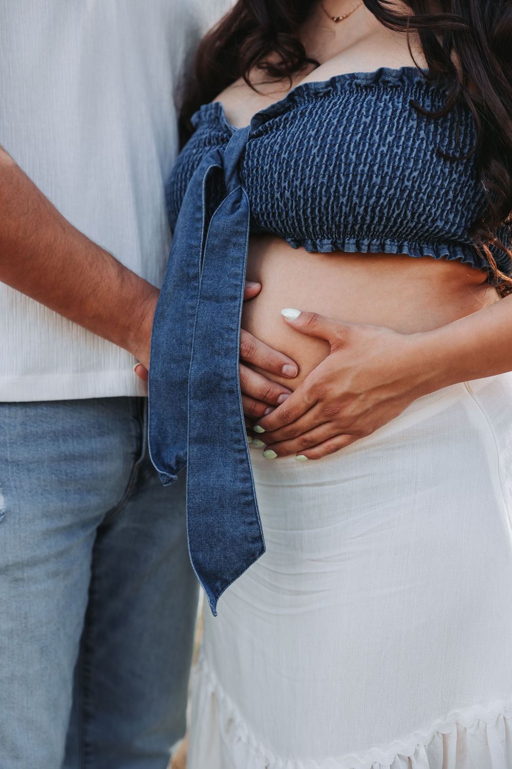 Pregnant woman in blue smocked crop top and white skirt with partner's hands cradling baby bump outdoors.