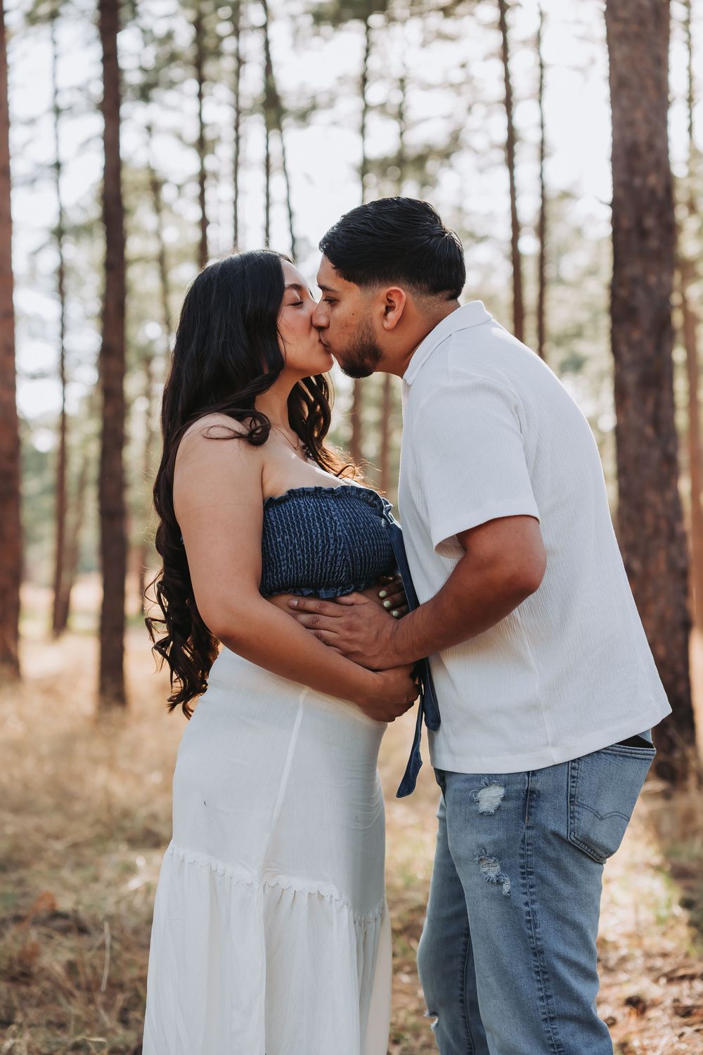 Couple sharing a kiss in a pine forest, woman in white dress with blue smocked top showing baby bump.