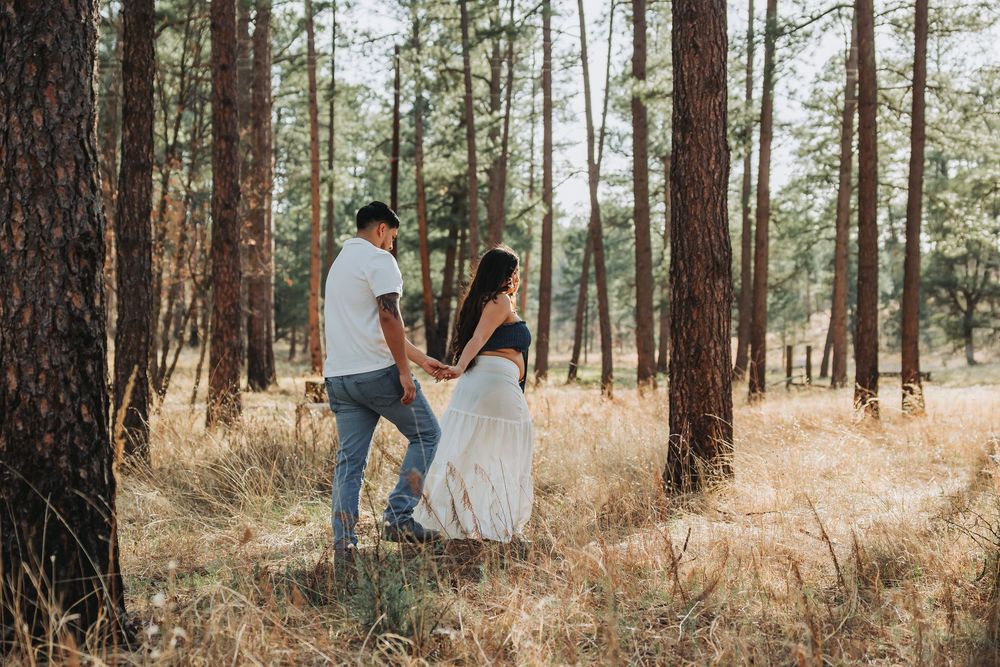 Couple holding hands walking through a sunlit pine forest, woman in white dress and man in casual attire.