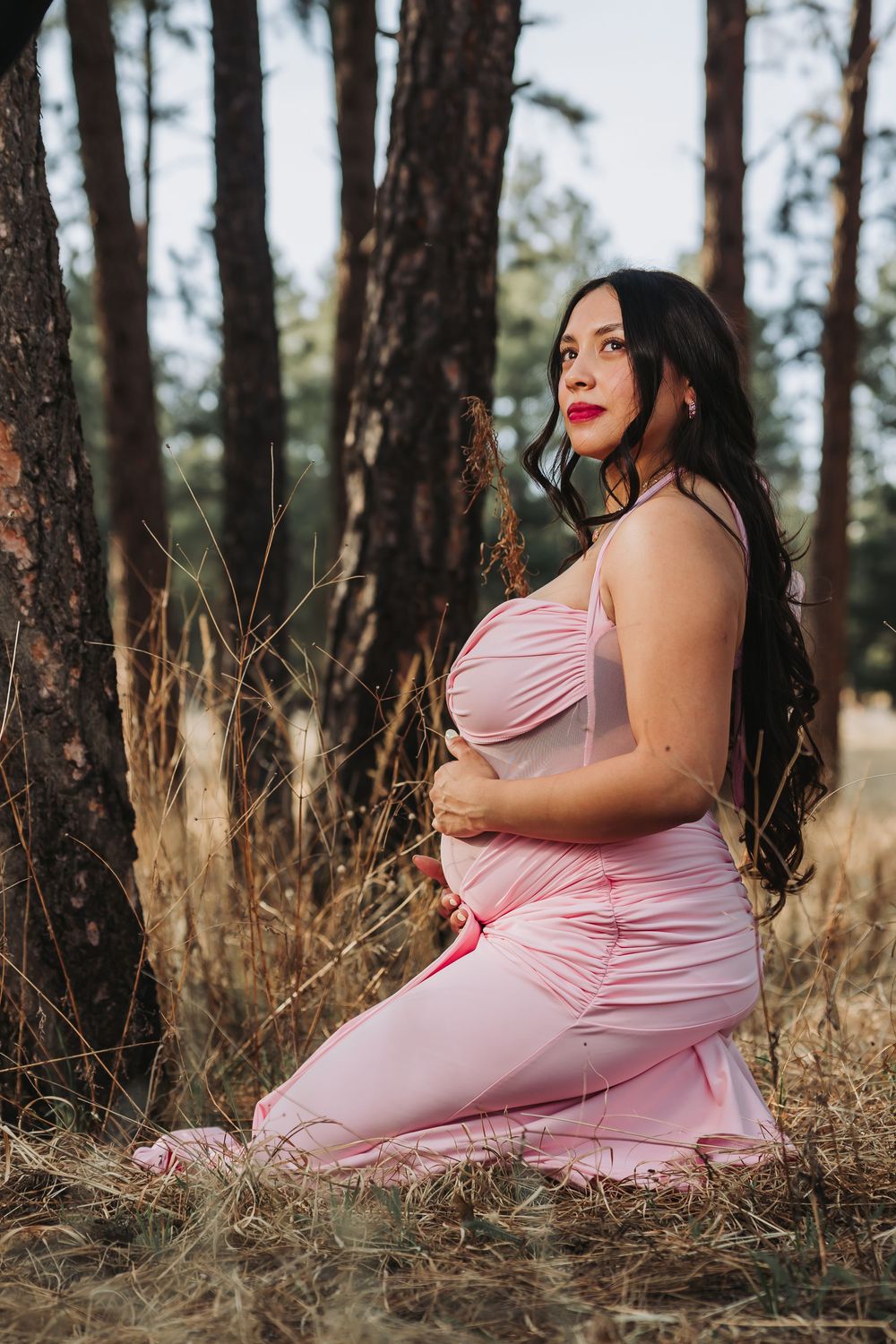 Pregnant woman in pink dress kneeling in forest, cradling baby bump among pine trees in a serene maternity photo.