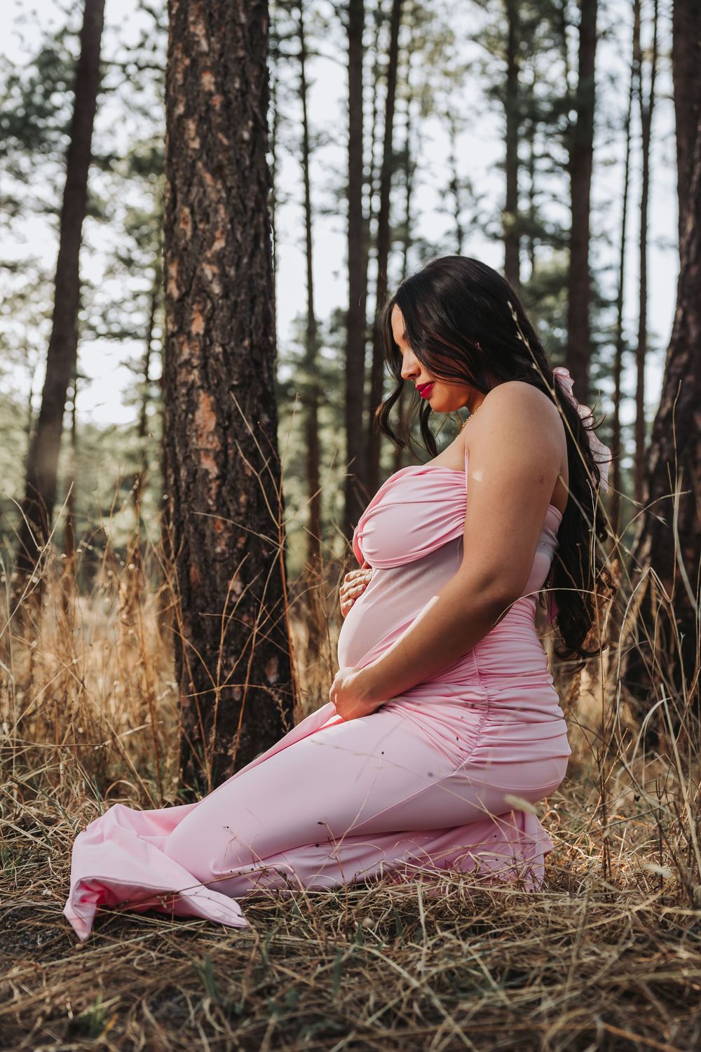 Pregnant woman in pink dress sitting peacefully among pine trees, cradling her baby bump in a serene forest setting.