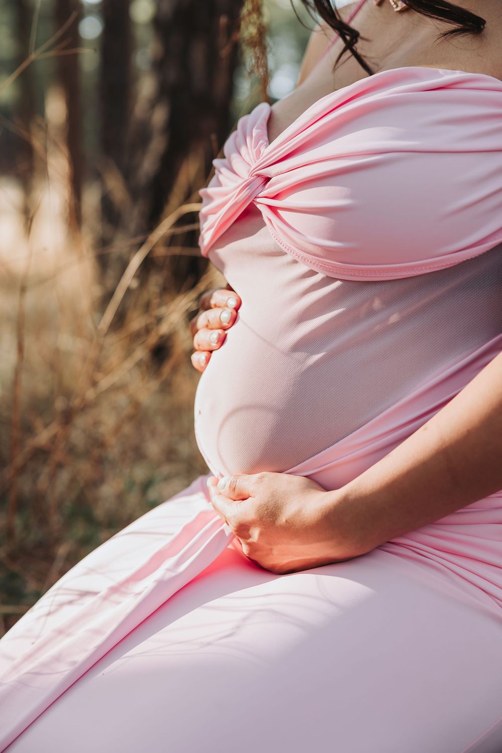 Pregnant woman in pink off-shoulder dress cradling baby bump outdoors in natural forest setting.