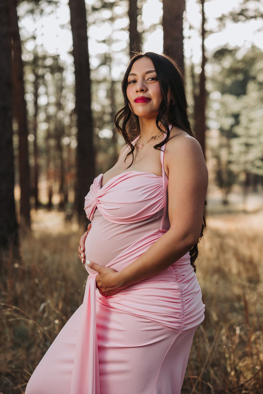Pregnant woman in pink ruched maternity gown poses gracefully in a sunlit forest setting.