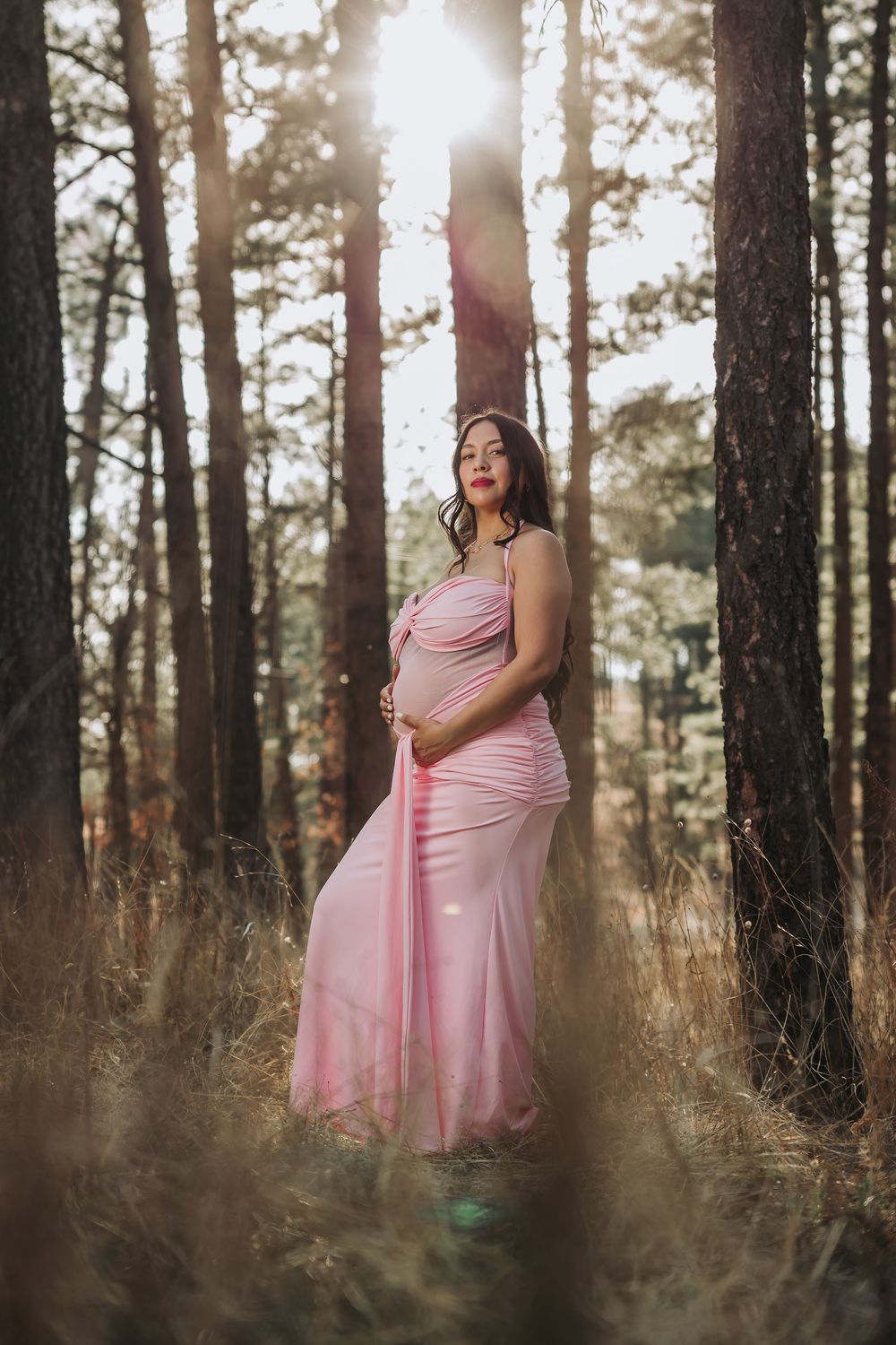 Pregnant woman in pink gown standing among tall pine trees with sunlight streaming through forest backdrop.