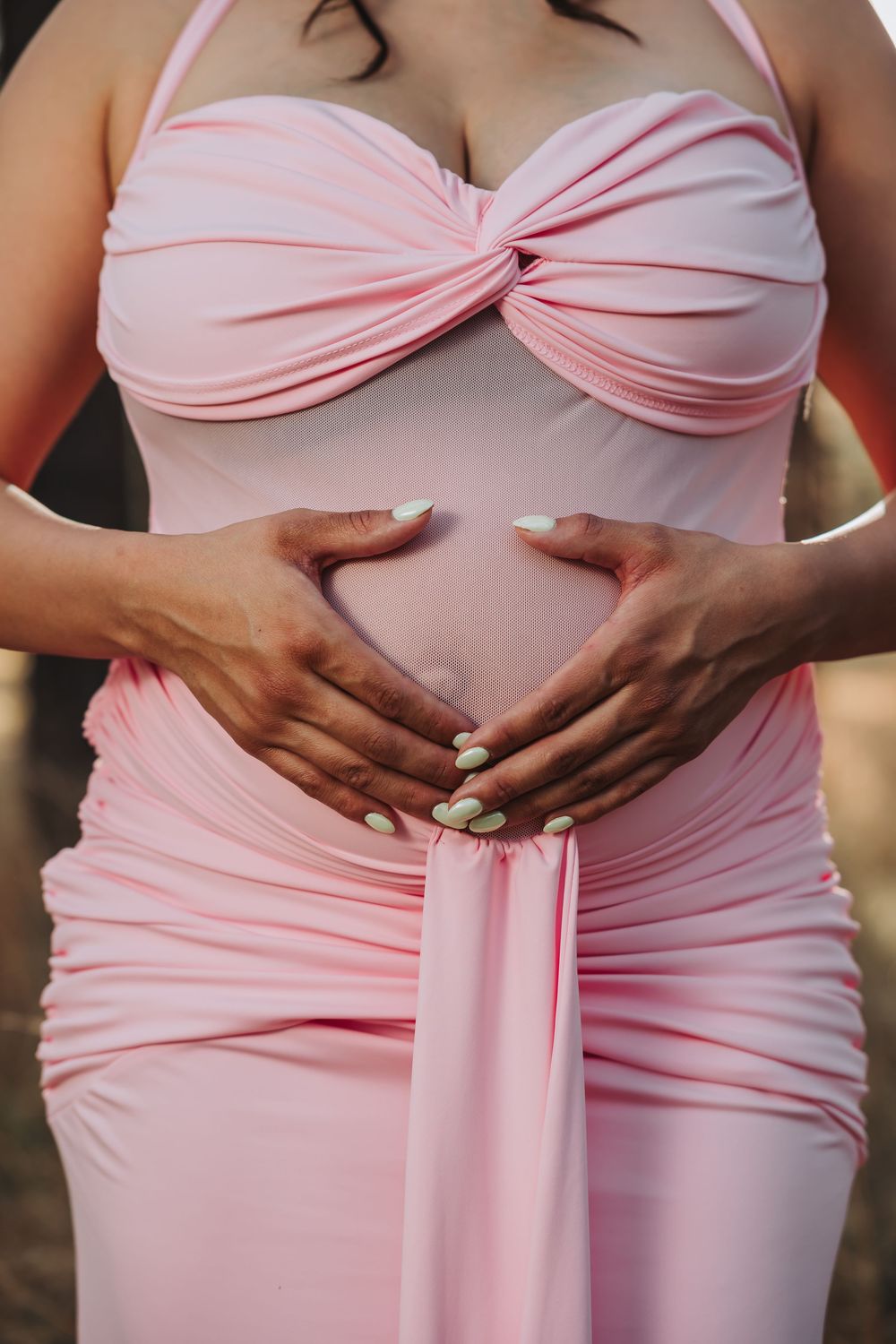 Pregnant woman in pink ruched maternity dress cradling baby bump with both hands outdoors.