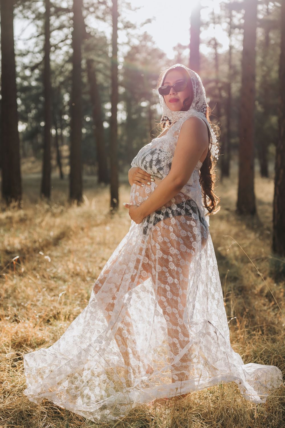 Pregnant woman in sheer lace dress and sunglasses cradling her baby bump in a sunlit forest.
