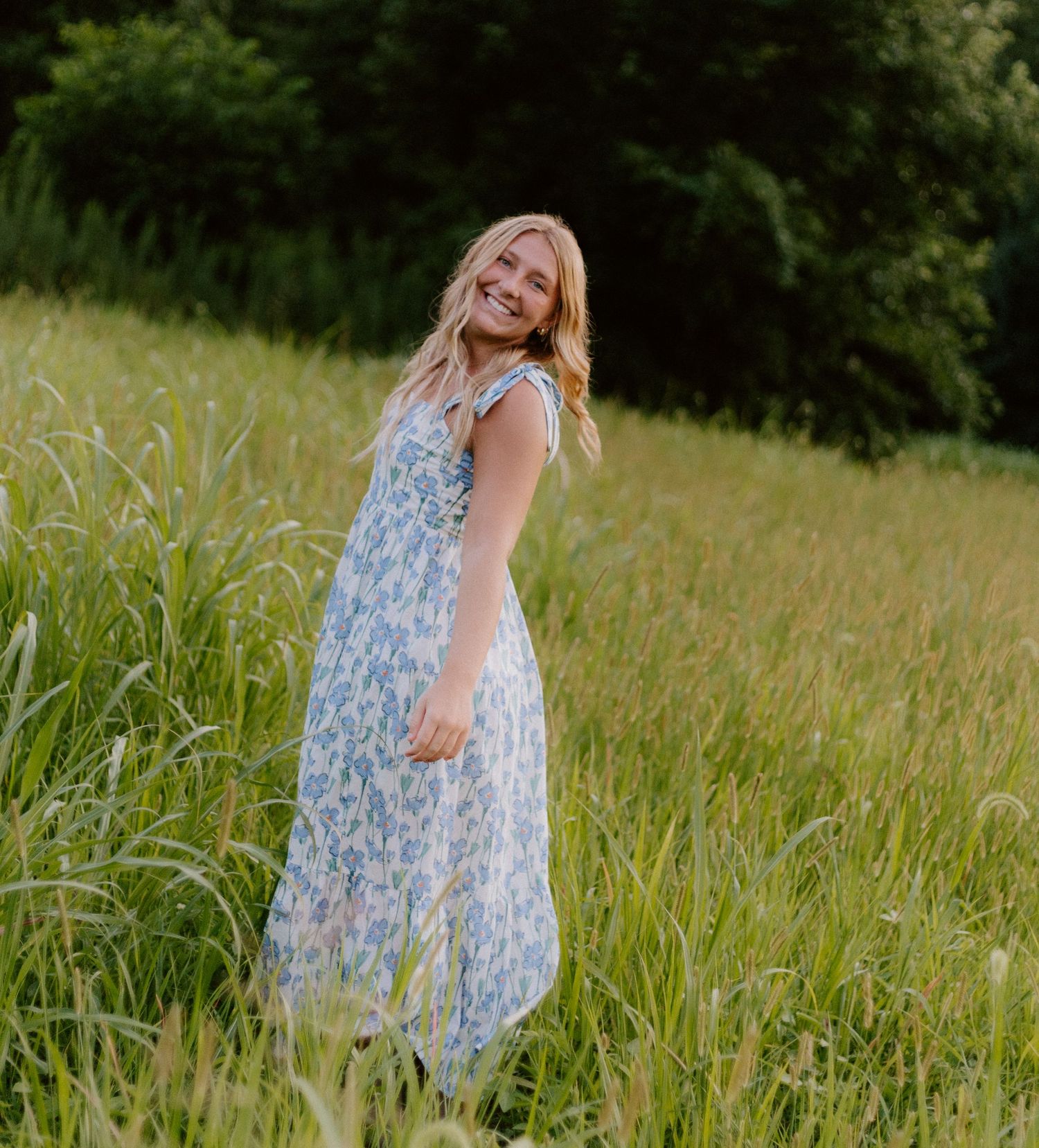 Smiling blonde woman in blue floral maxi dress standing in a lush green meadow.