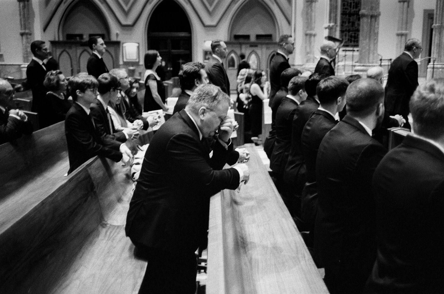 Black and white photo of elegantly dressed guests praying in a grand Midwest Basilica during a formal wedding ceremony.