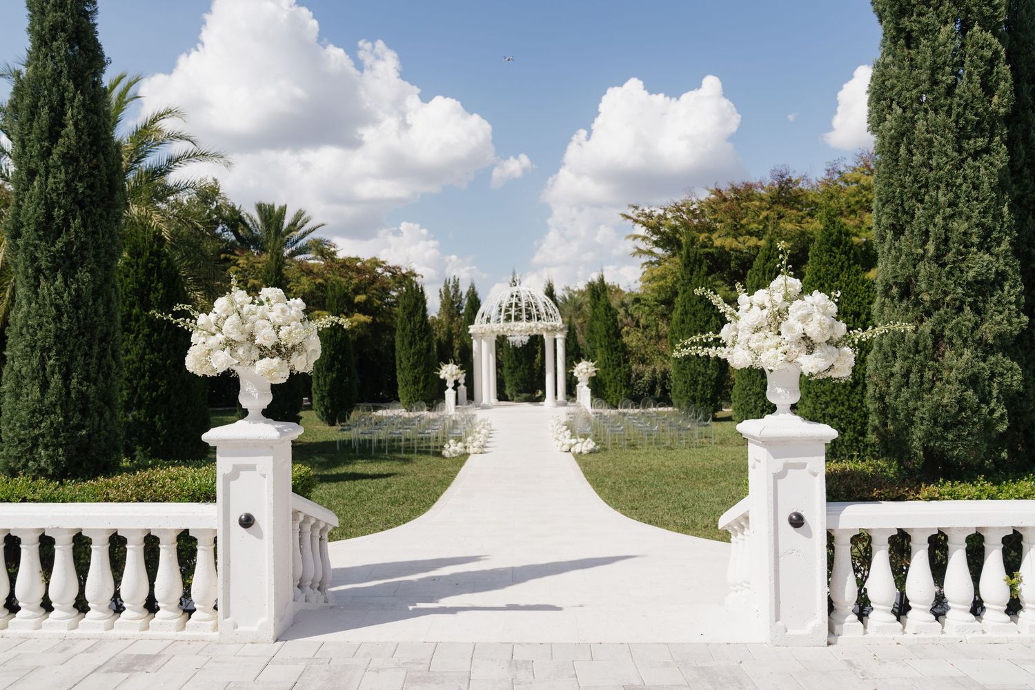 Elegant white wedding ceremony aisle with floral urns, cypress trees, and a gazebo in a manicured garden.
