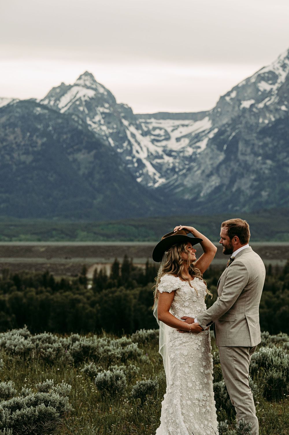 Bride in lace gown and cowboy hat with groom in gray suit, embracing before snow-capped Grand Teton mountains.