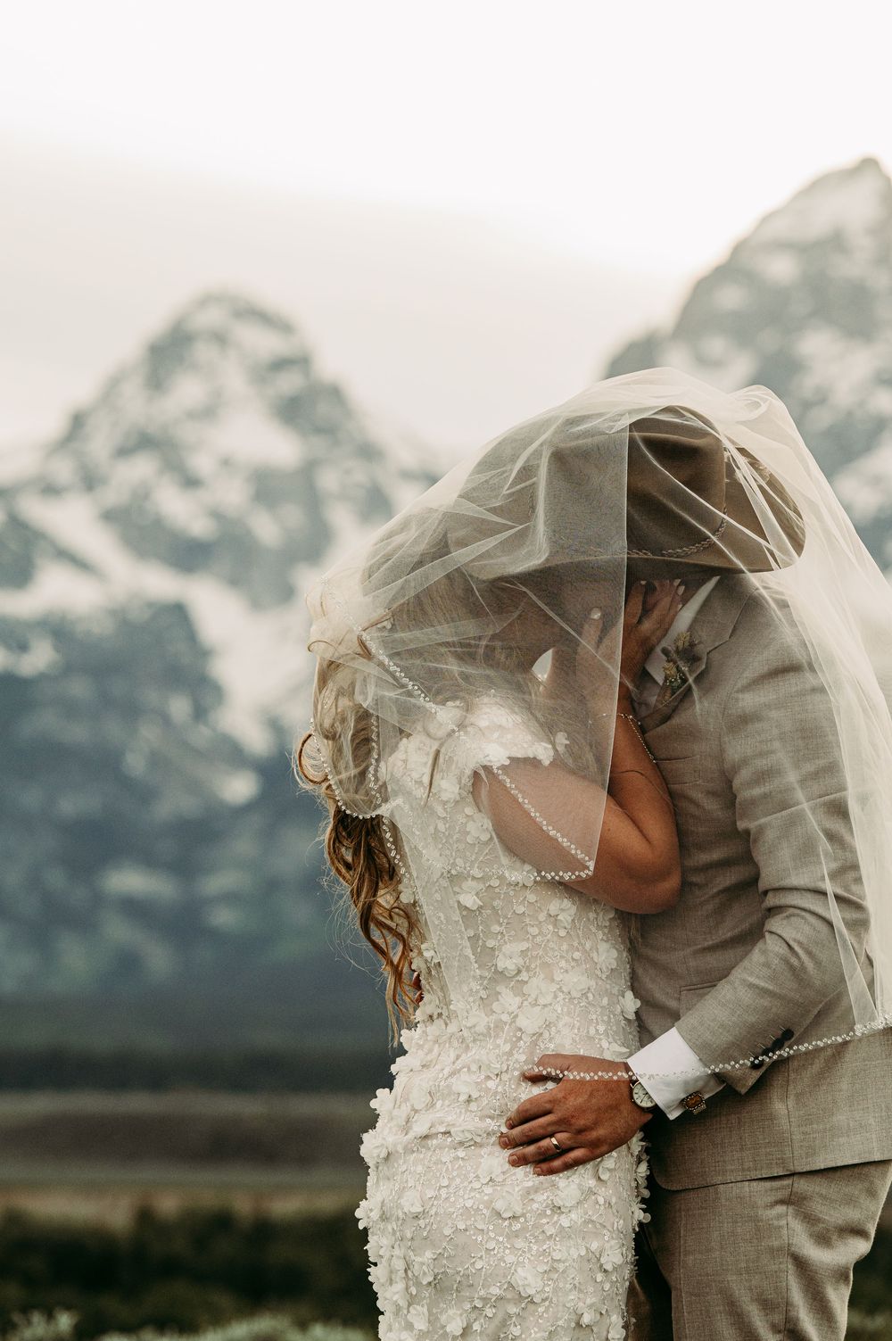 Bride and groom share a kiss under a veil with snow-capped Grand Teton mountains in the background.