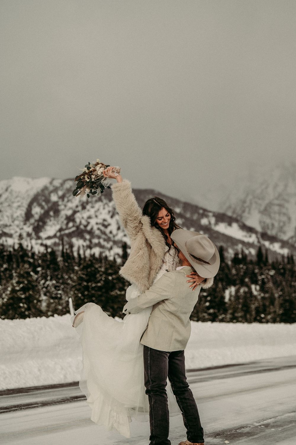 Couple celebrating winter wedding in snowy mountains, bride holding bouquet aloft while groom lifts her, wearing fur coat and cowboy hat.