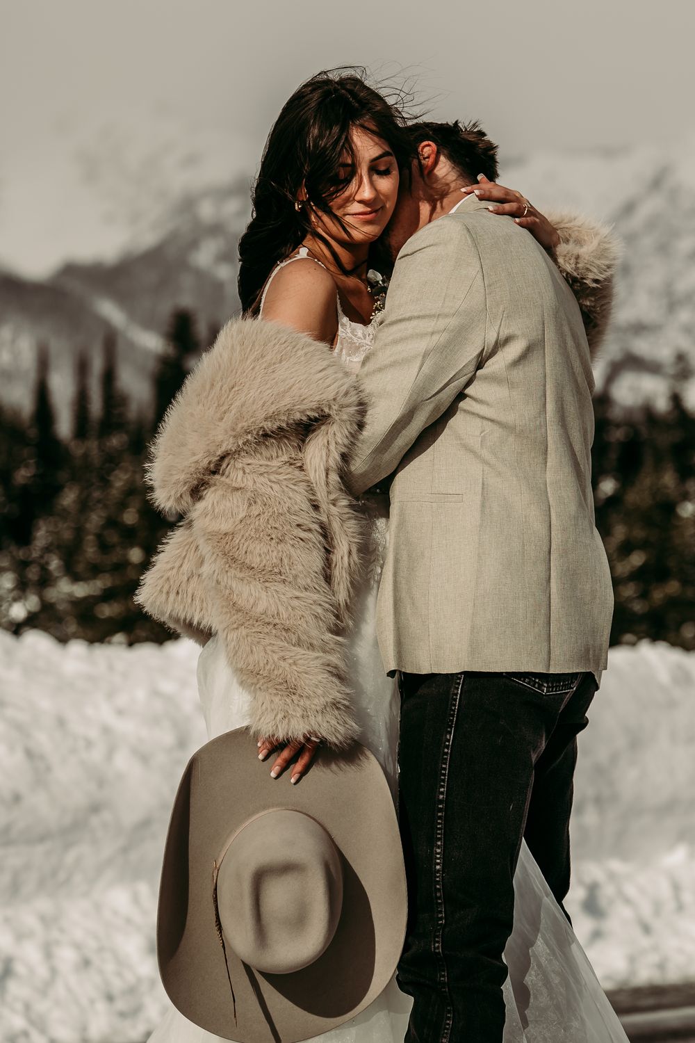 Bride in fur coat holding cowboy hat embraces groom in snowy mountain landscape during winter wedding elopement.