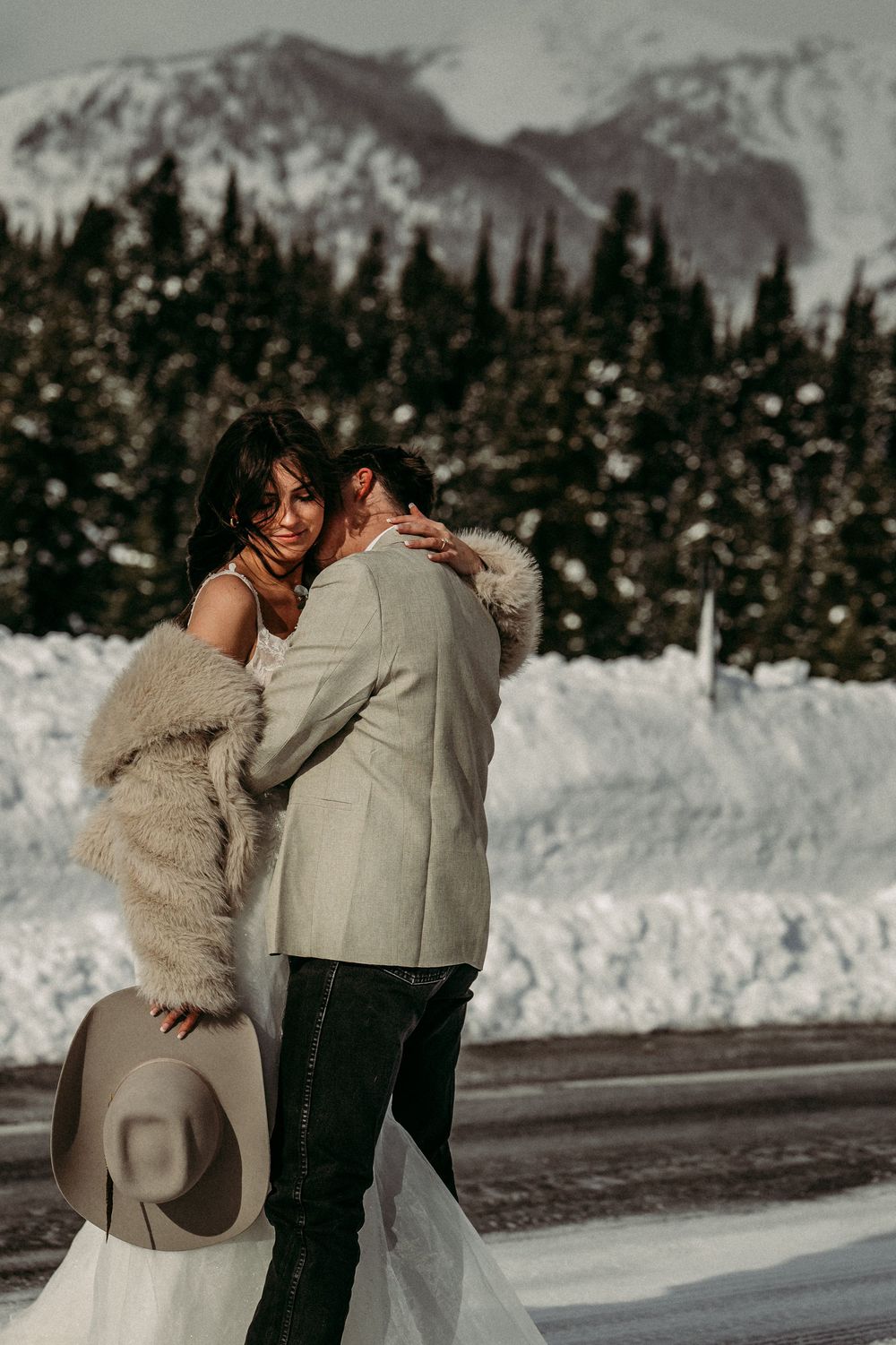Bride in fur stole and groom in cream suit embrace in snowy mountain landscape during winter wedding elopement.