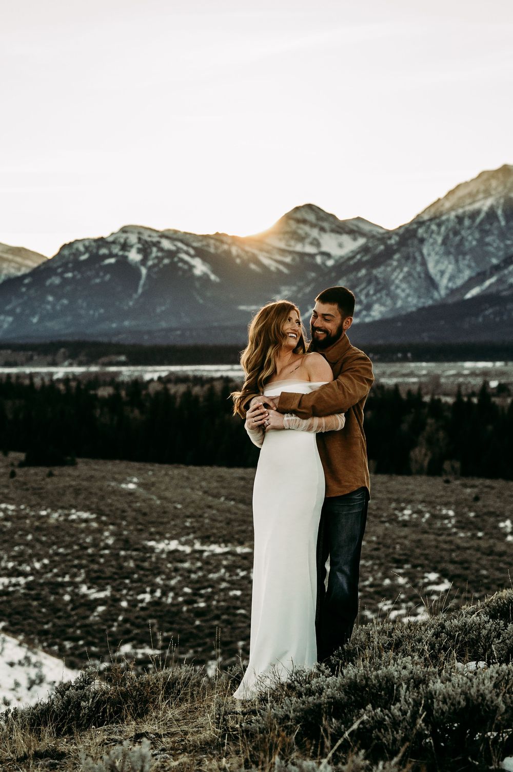 Couple embracing at golden hour with snow-capped mountains behind them, woman in white gown and man in brown jacket.