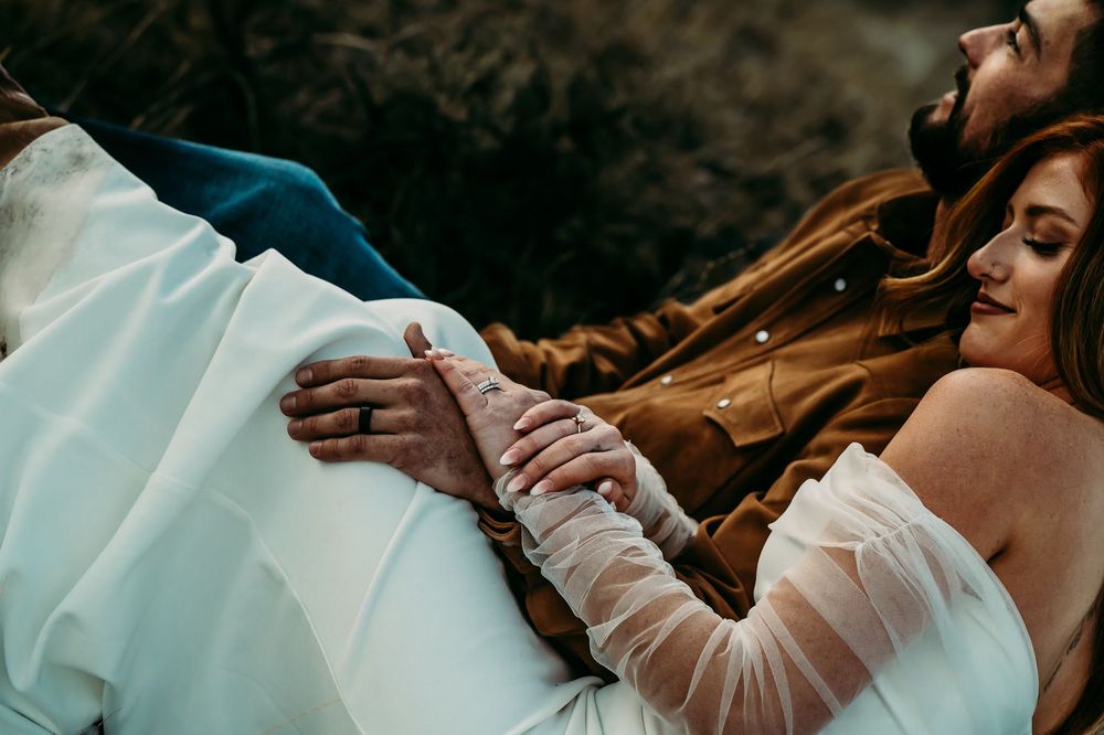 Couple holding hands tenderly, woman in white dress and man in brown jacket, sharing a romantic moment outdoors.