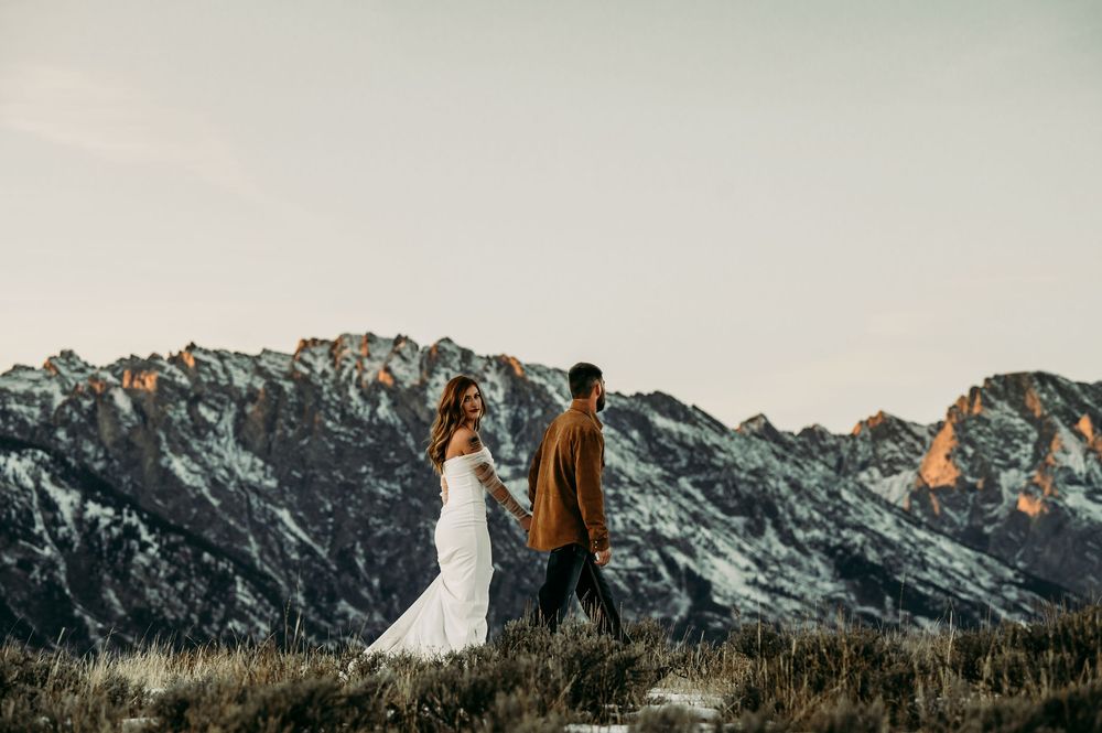 Couple in wedding attire holding hands in a mountain meadow with snow-capped peaks at sunset.