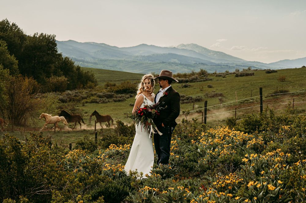 Bride and groom embrace in wildflower field with horses and mountain backdrop at rustic outdoor wedding.